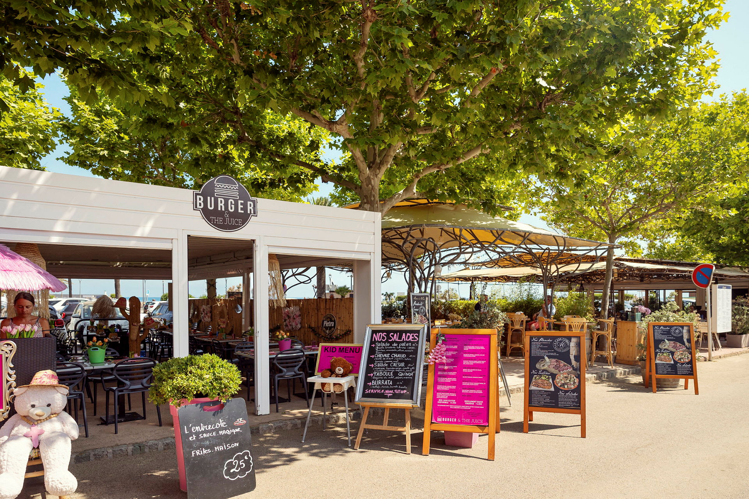 Utomhusrestaurang med skyltar och växtligheter. Bord och stolar står under trädkronor och parasoller. (AI-genererad text)