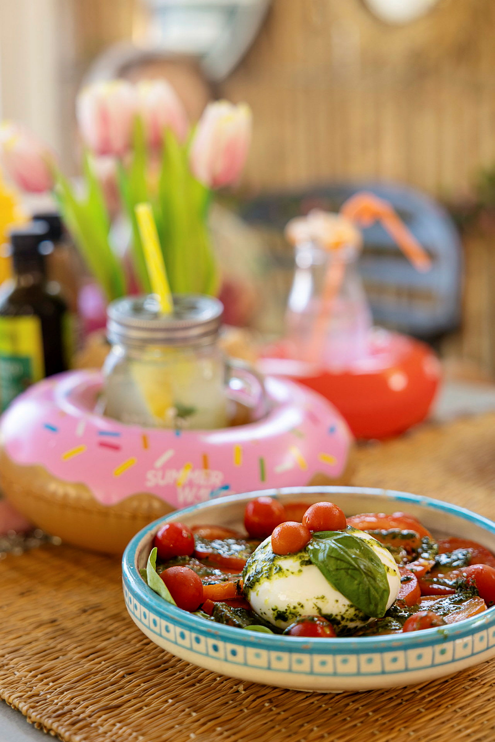 En maträtt med buffelmozzarella, tomater och basilika samt drycker med dekorationer. Bakgrunden visar tulpaner och andra prydnadsobjekt. (AI-genererad text)