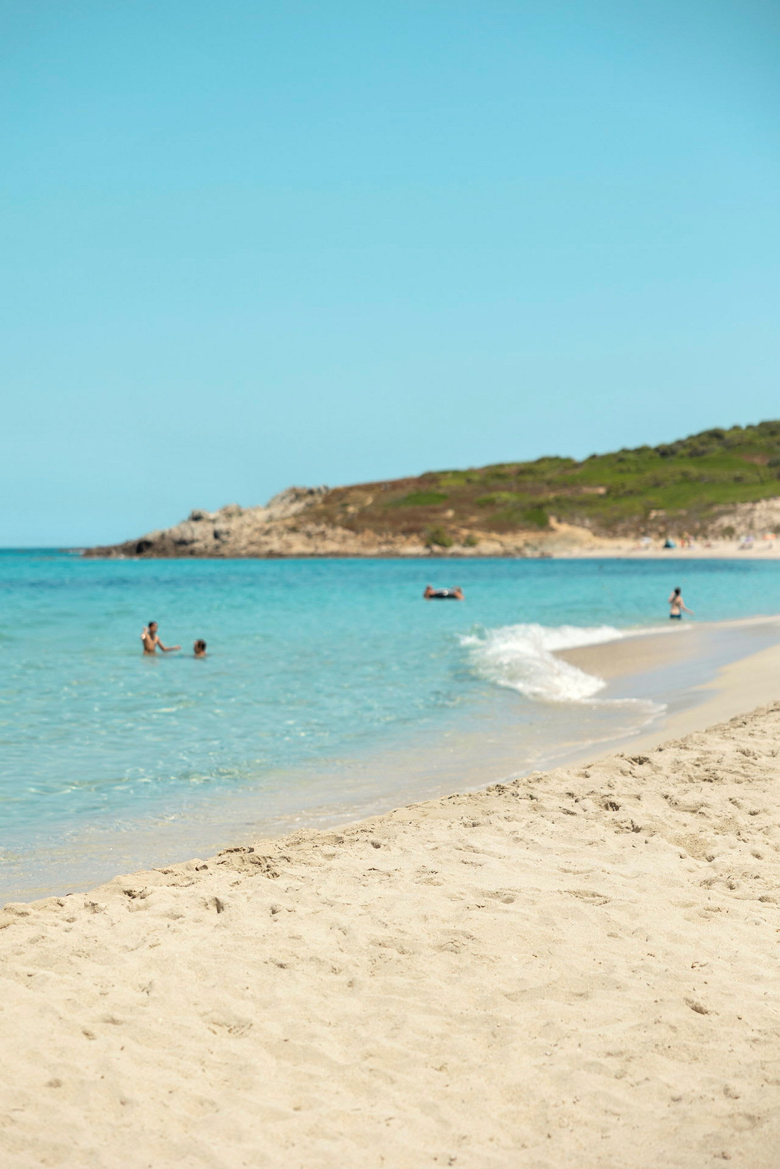 Sandstrand med hav och människor som badar, samt en grönskande kulle i bakgrunden. (AI-genererad text)