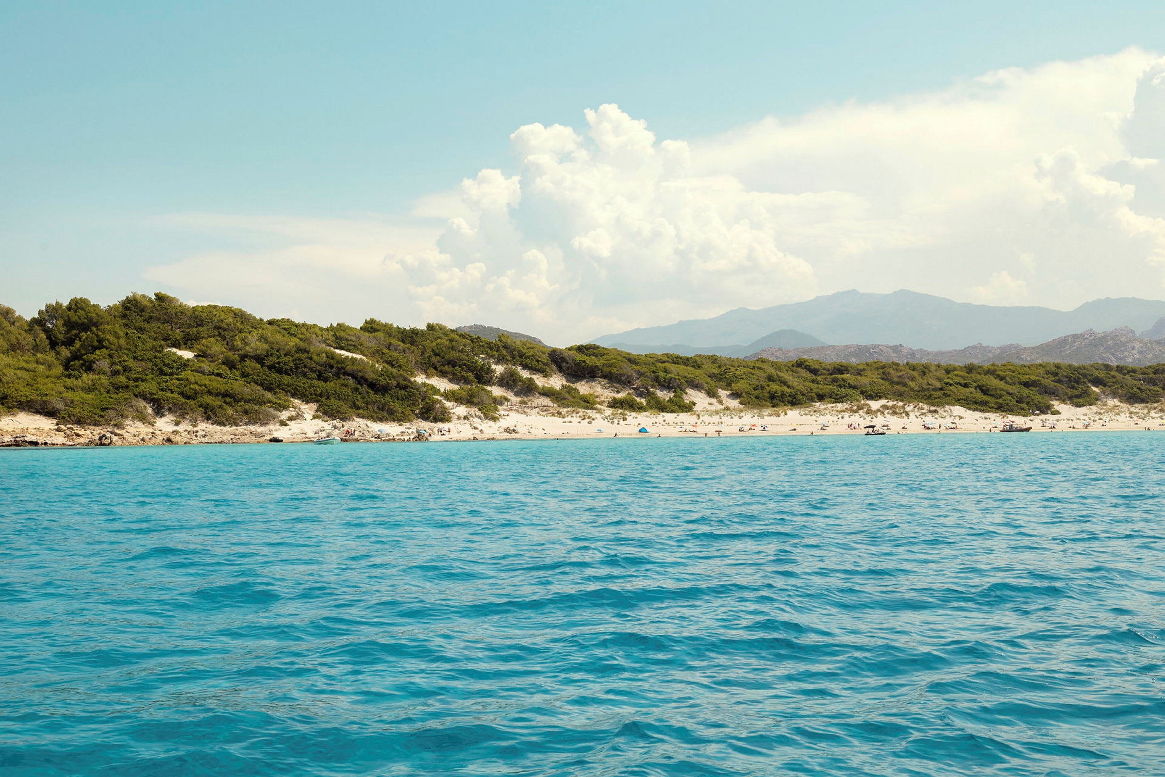 En strand med turkost vatten, omgiven av grön vegetation och berg i bakgrunden. (AI-genererad text)