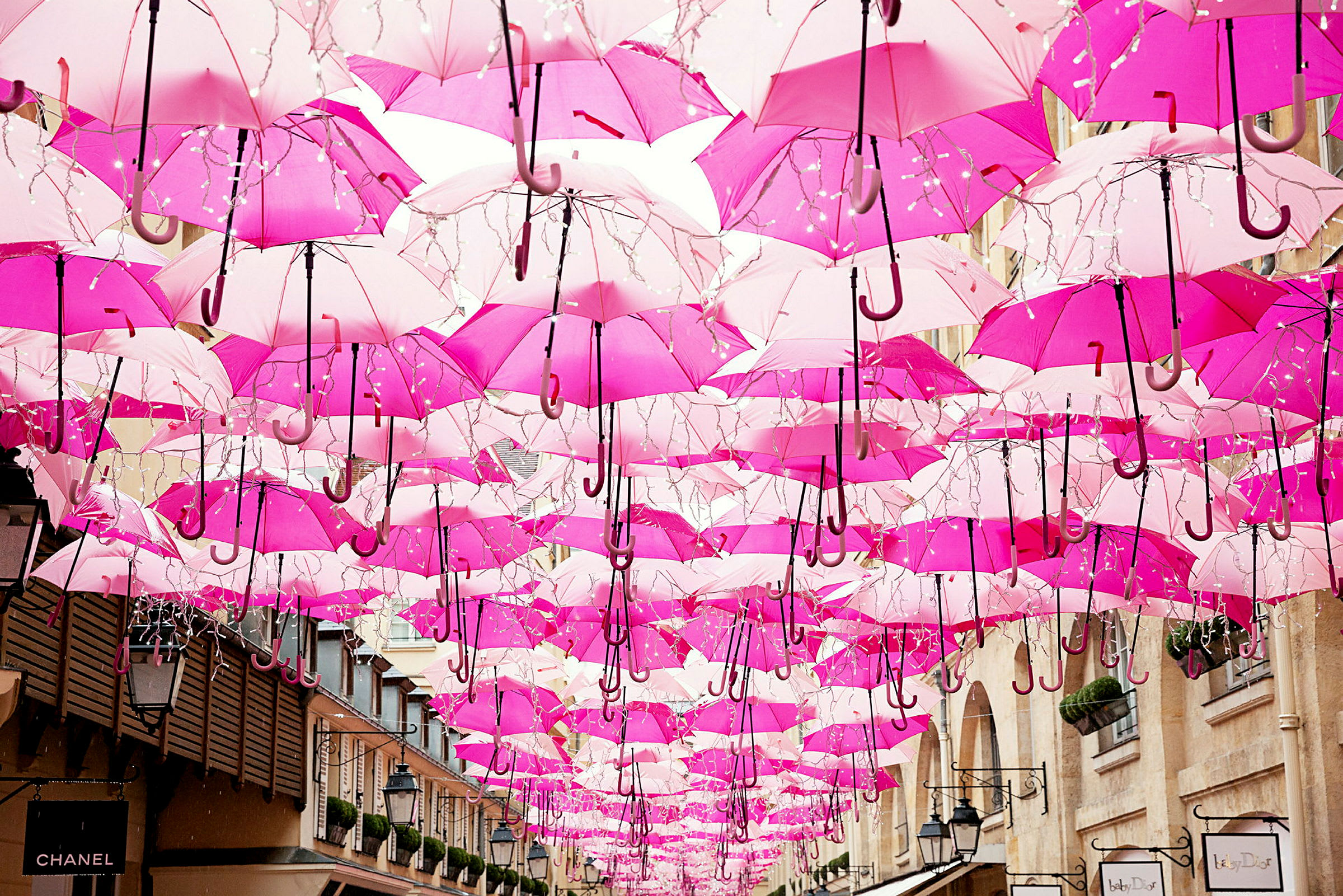 Flera rosa paraplyer hänger uppsatta i luften mellan byggnader. (AI-genererad text)