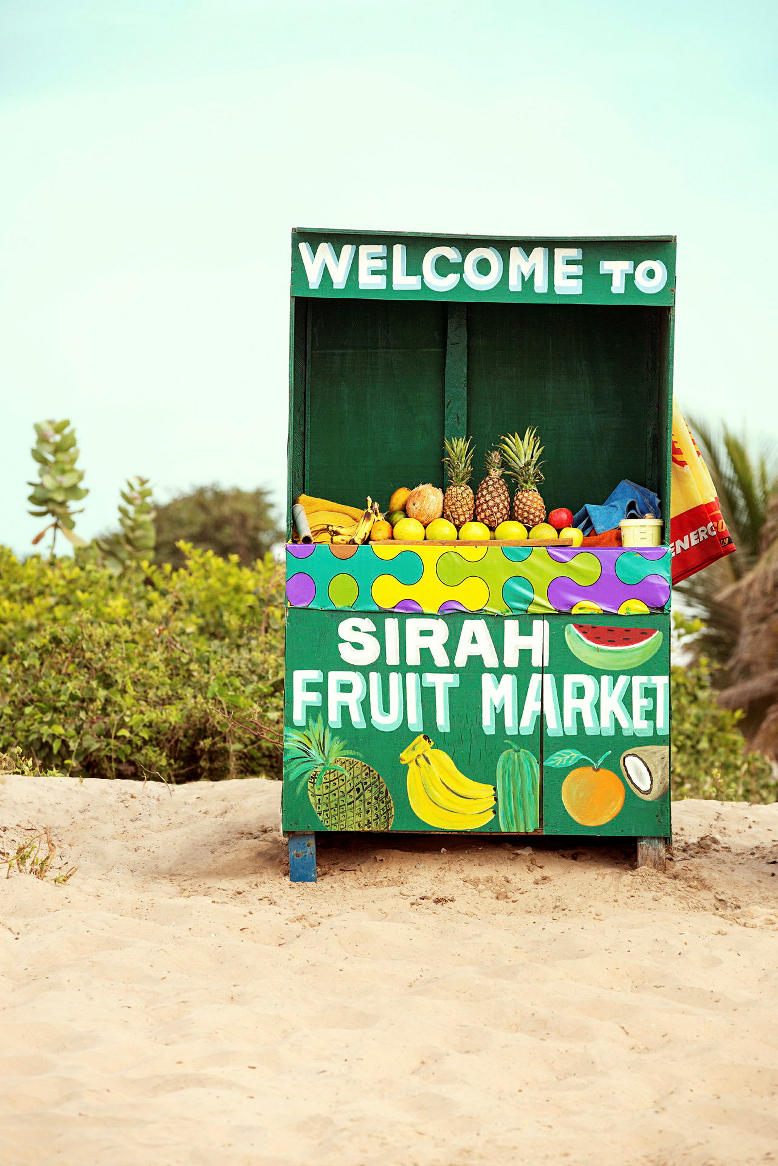 Ett fruktstånd med texten "Sirah Fruit Market" erbjuder en mängd olika frukter, inklusive ananas, bananer och citrusfrukter. Ståndet står på sand med vegetation i bakgrunden. (AI-genererad text)