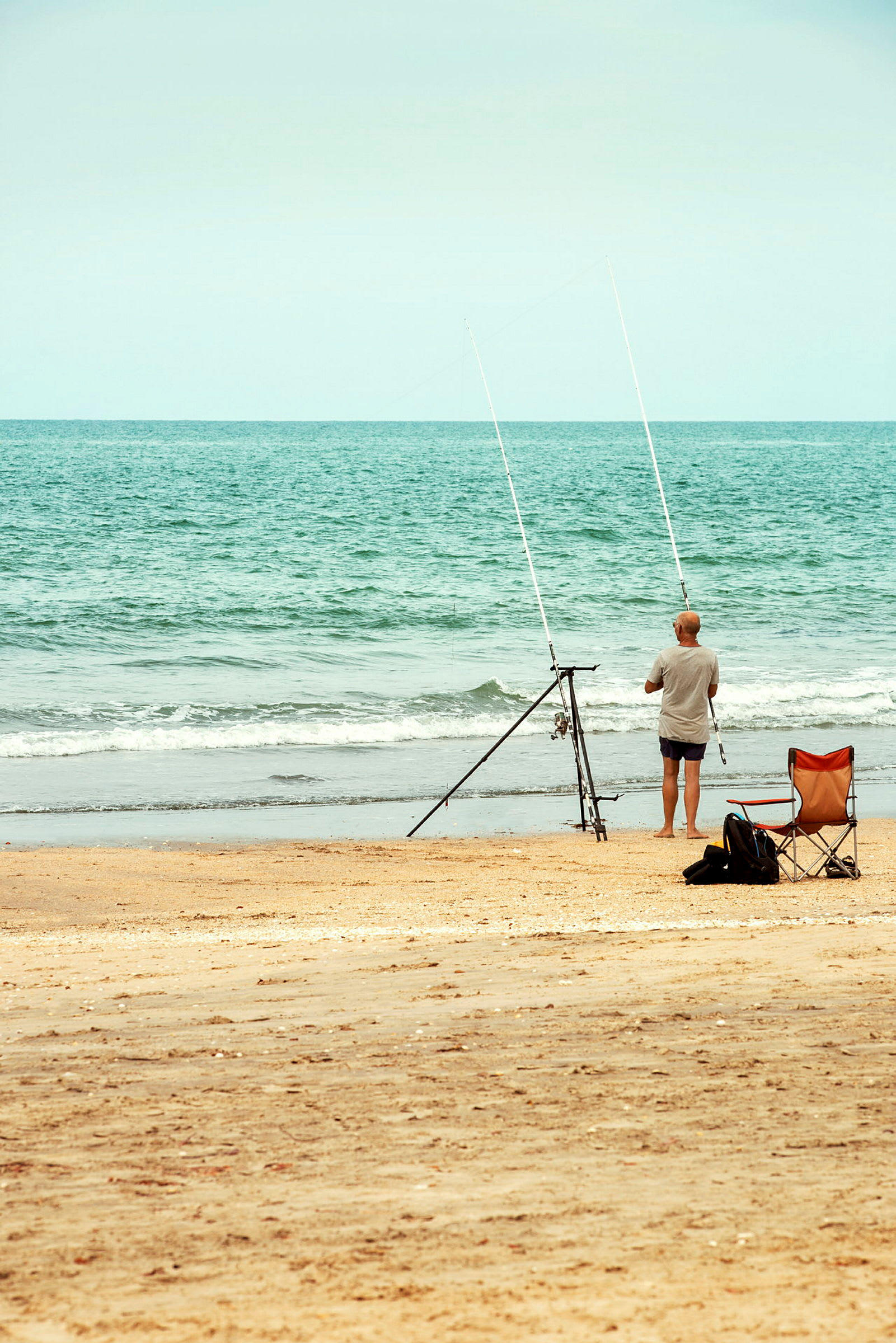 En person står vid strandkanten med två fiskespön på ett ställ. En stol står i sanden intill. (AI-genererad text)