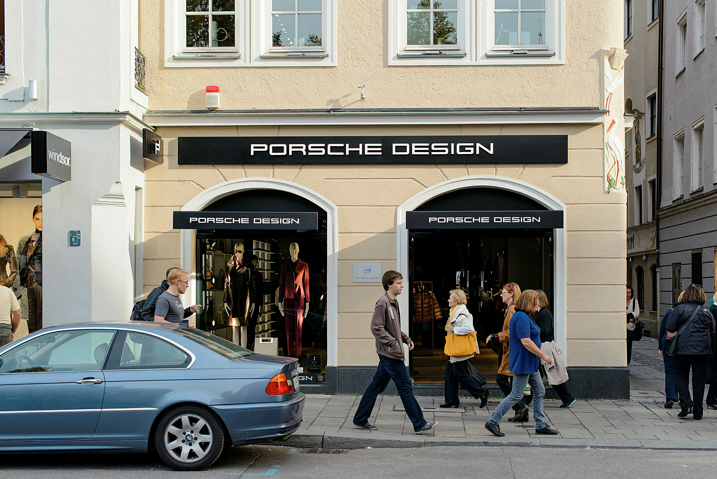 Entré till en butik med Porsche Design-logotyp, med skyltfönster och människor som går på trottoaren utanför. (AI-genererad text)