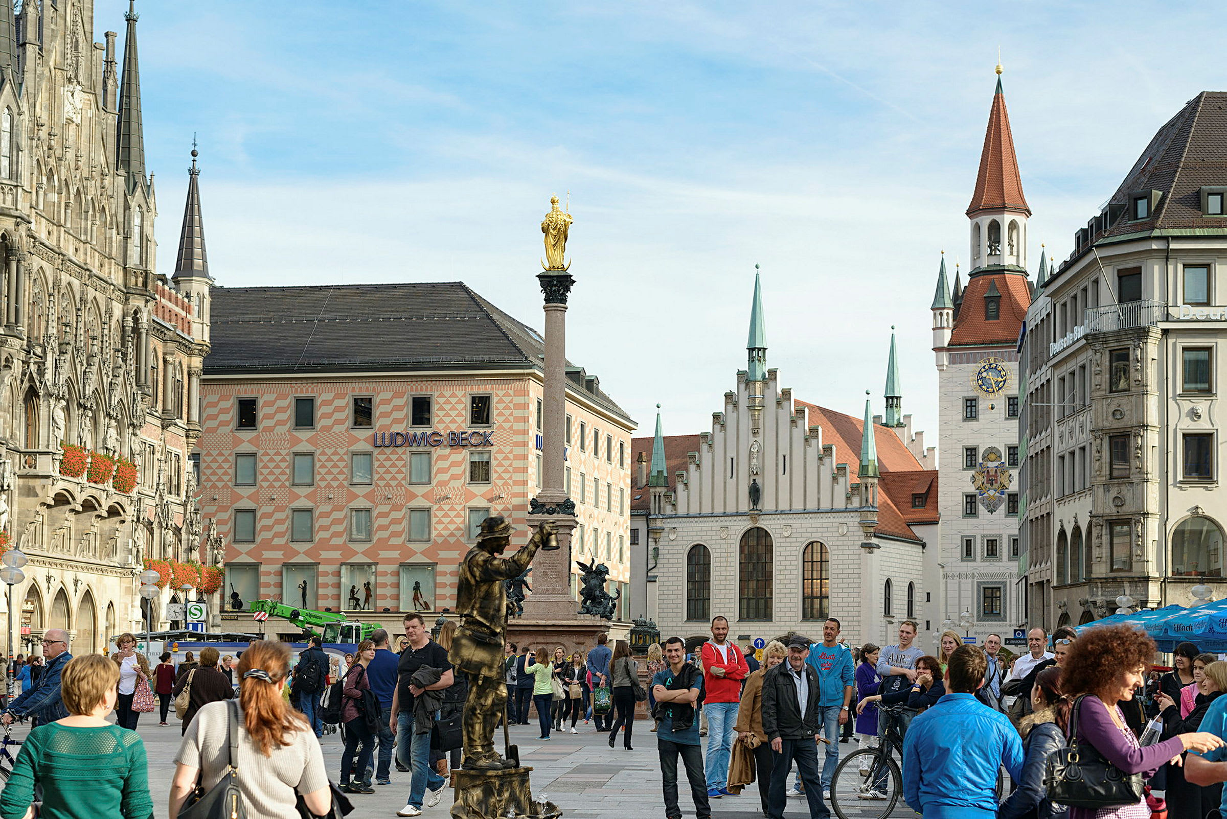 Ett livligt torg med historiska byggnader, en staty med en gyllene topp och många gående människor. (AI-genererad text)