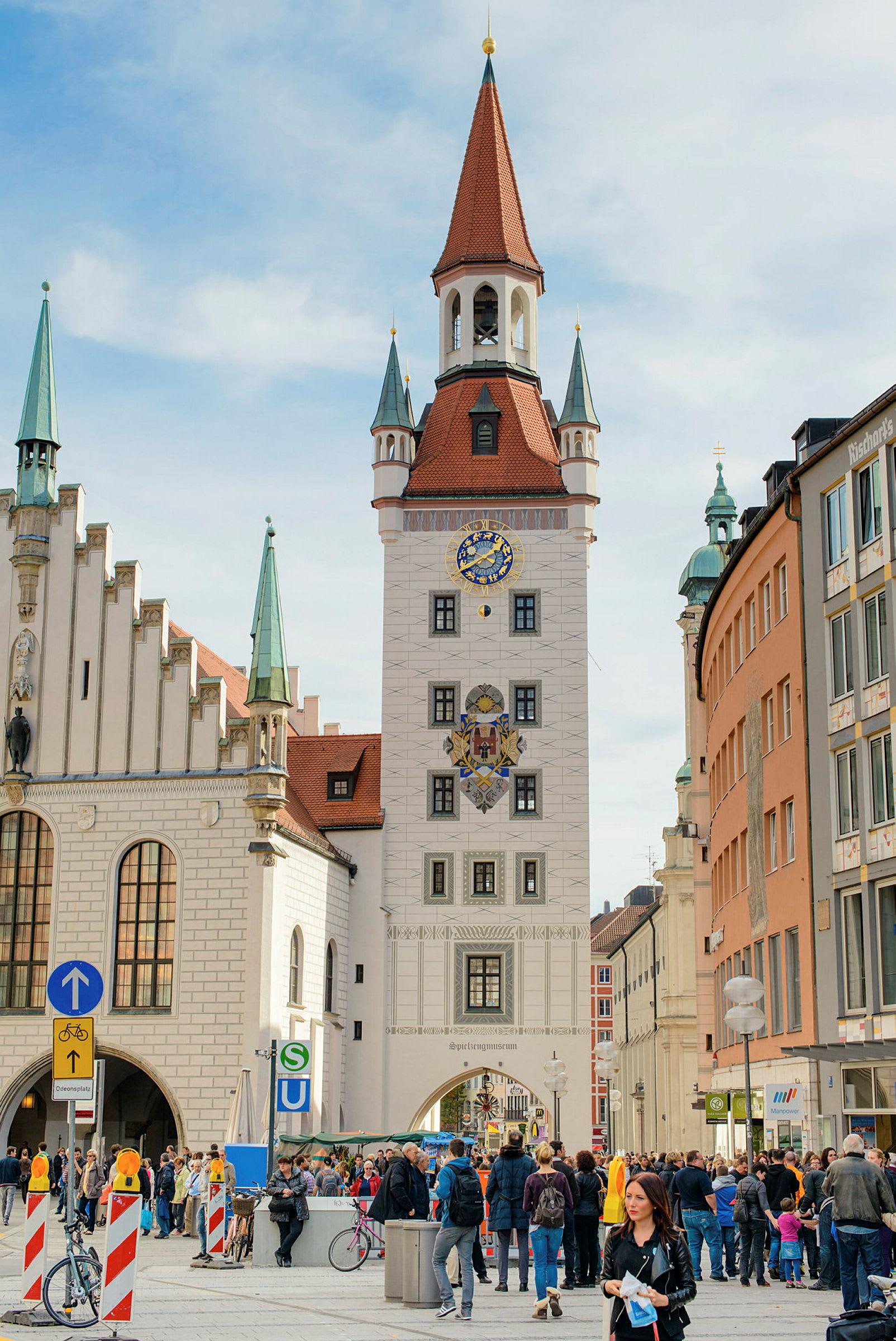 Bild av ett torg med ett högt torn i bakgrunden. Tornet har en klocka och detaljerad arkitektur, omgärdat av byggnader. Människor är samlade på torget, några står och några går. (AI-genererad text)