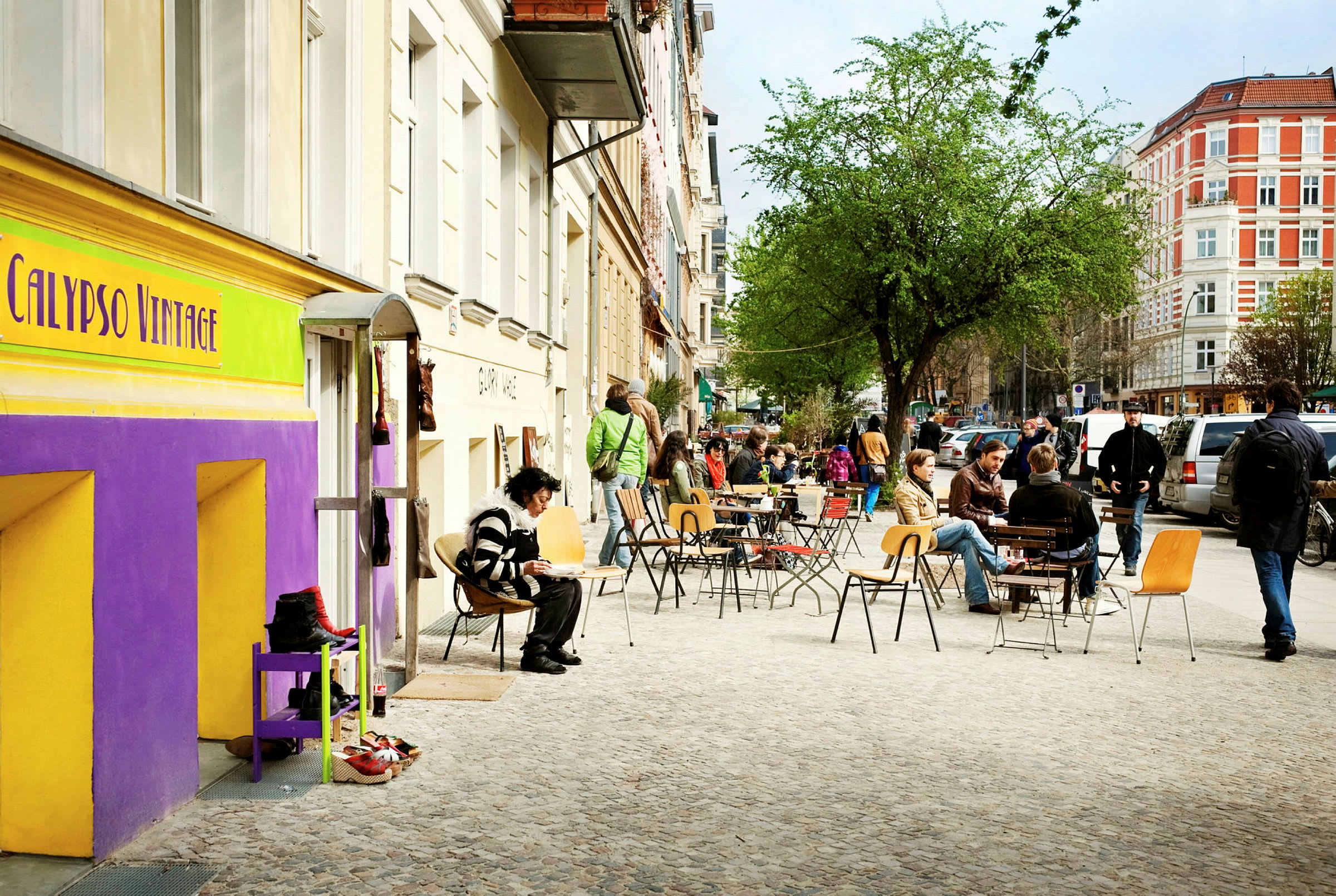 Gatan kantas av äldre byggnader med cafébord och sittplatser utomhus, där människor sitter och njuter av dagen. (AI-genererad text)