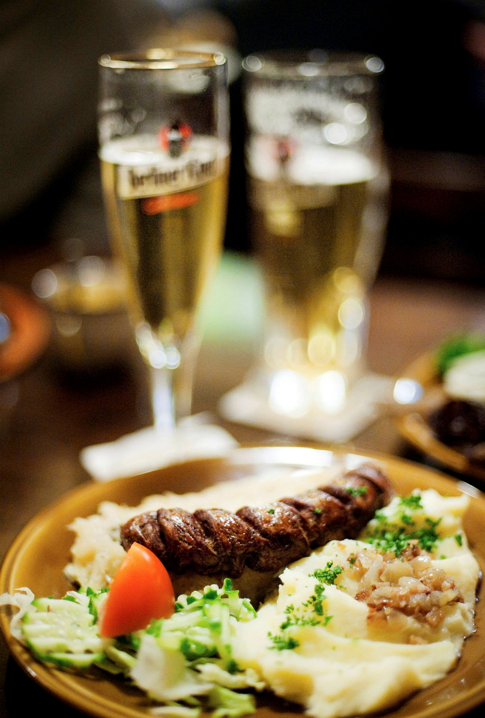 Tallrik med mat: köttkorv, potatismos med garnering, grönsaker och en tomatklyfta. I bakgrunden syns två ölglas. (AI-genererad text)