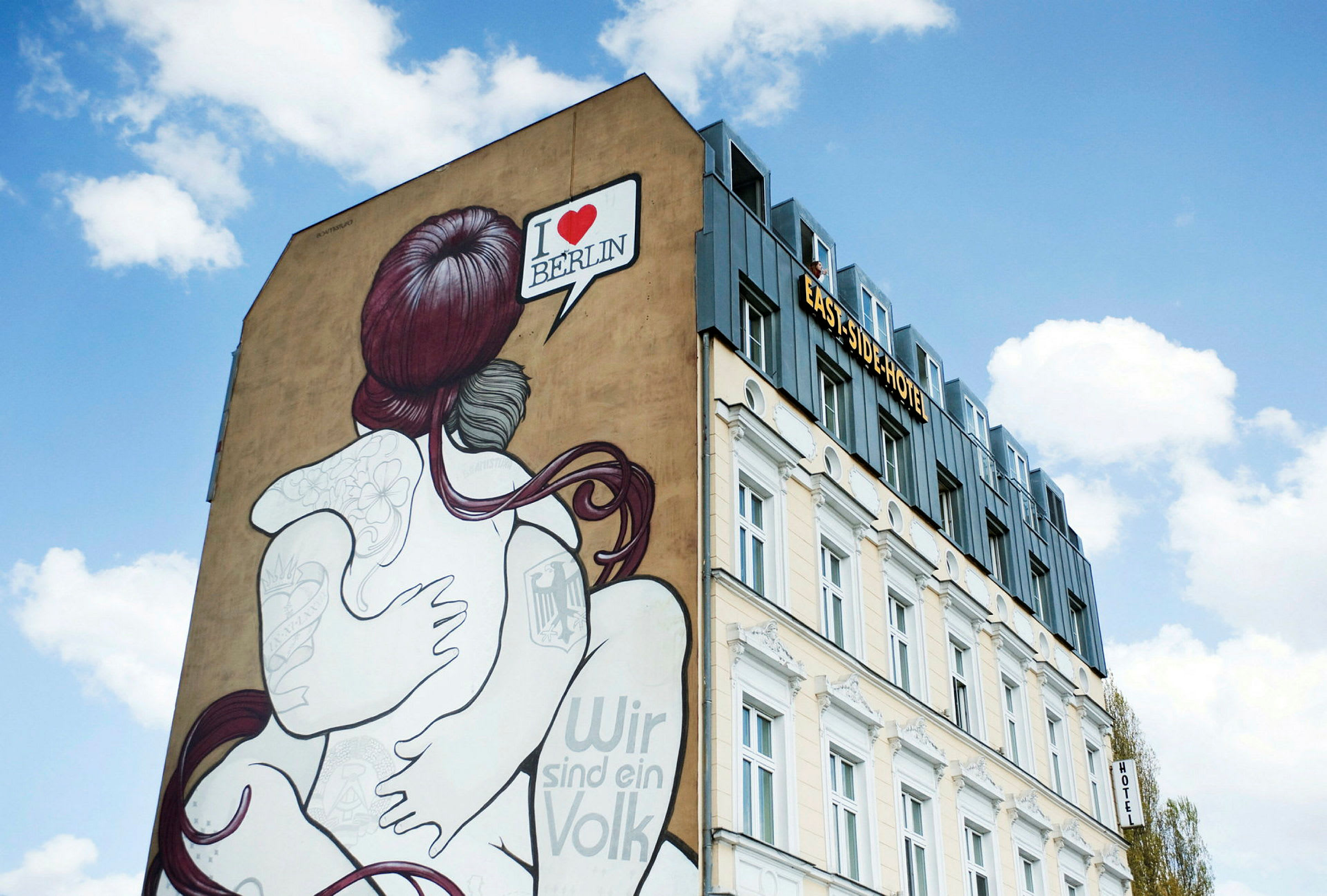En byggnad med en muralmålning av ett par som omfamnar varandra. Texten "I ♥ BERLIN" syns i ett pratbubbla. Himmelen är klar med vita moln. (AI-genererad text)