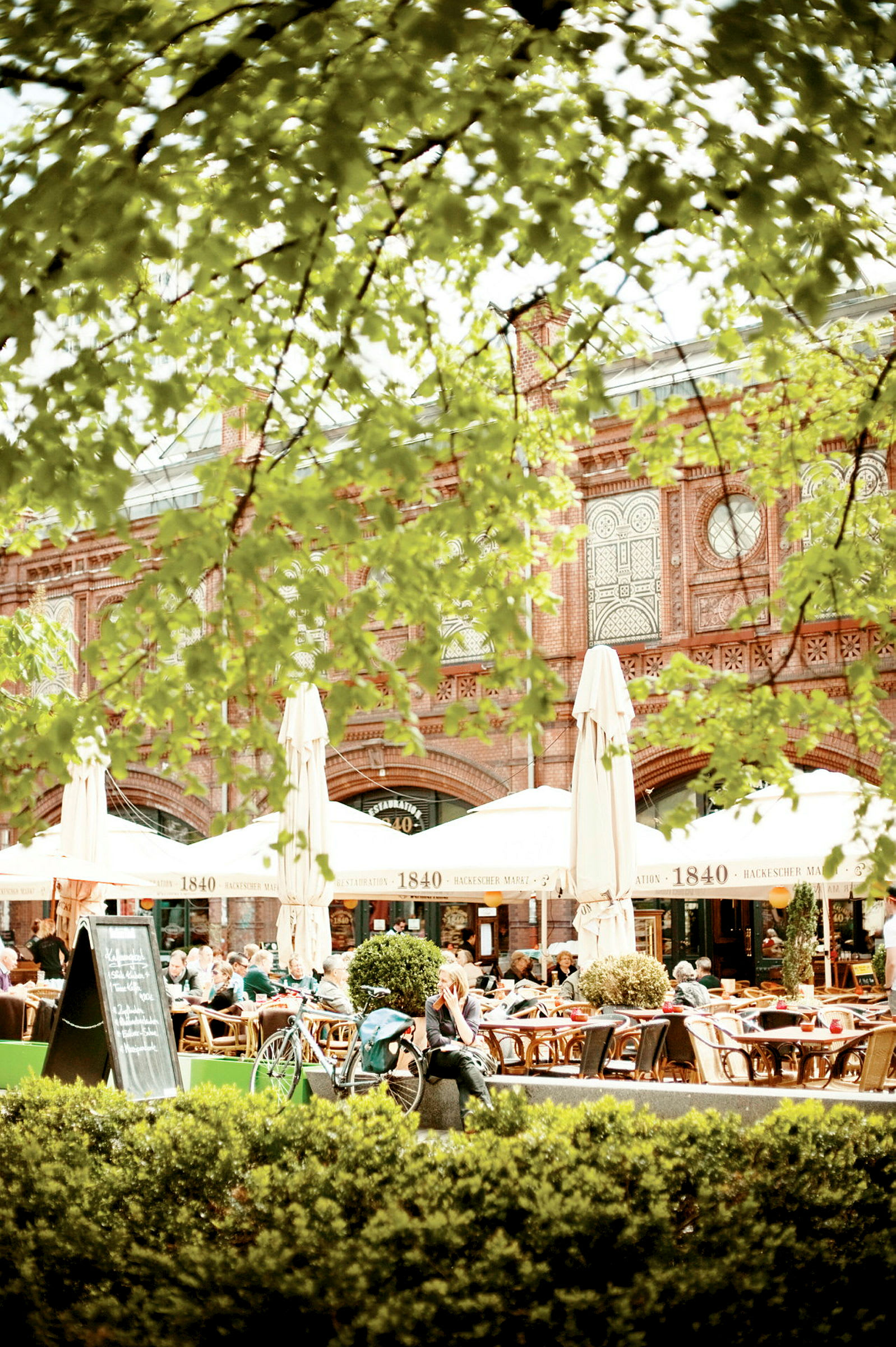 Ett kafé med uteservering under stora parasoller, omgivet av grönska och med en tegelbyggnad i bakgrunden. (AI-genererad text)