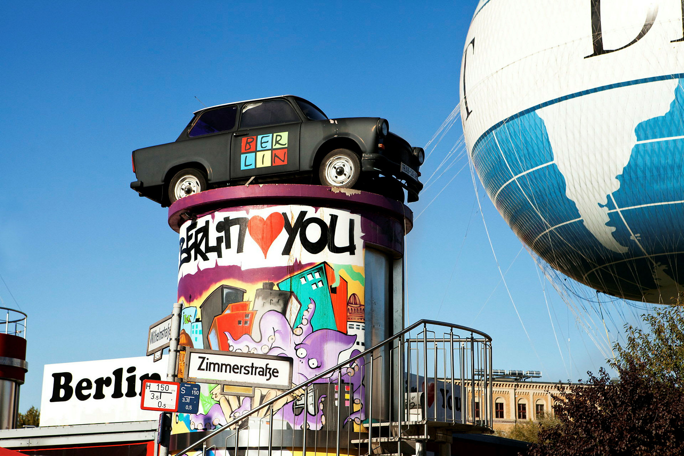 En vy av en svart bil placerad ovanpå en färgglad graffitimålad pelare bredvid en luftballong och en skylt med texten Zimmerstraße. (AI-genererad text)