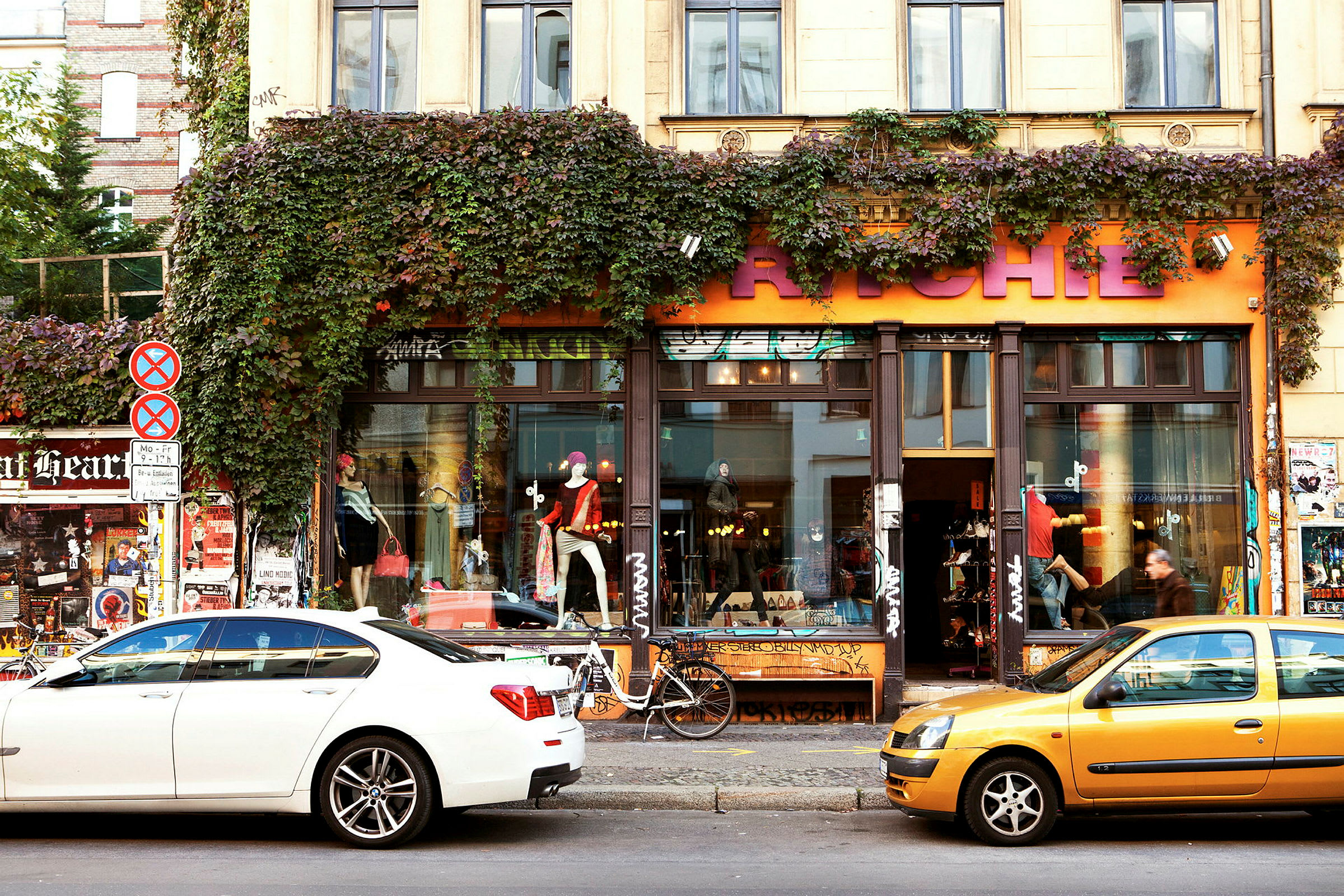 En byggnad med en butik har ett orangefärgat skylt med stora bokstäver, omgiven av gröna växter som klättrar längs fasaden. Bilar står parkerade framför butiken. (AI-genererad text)