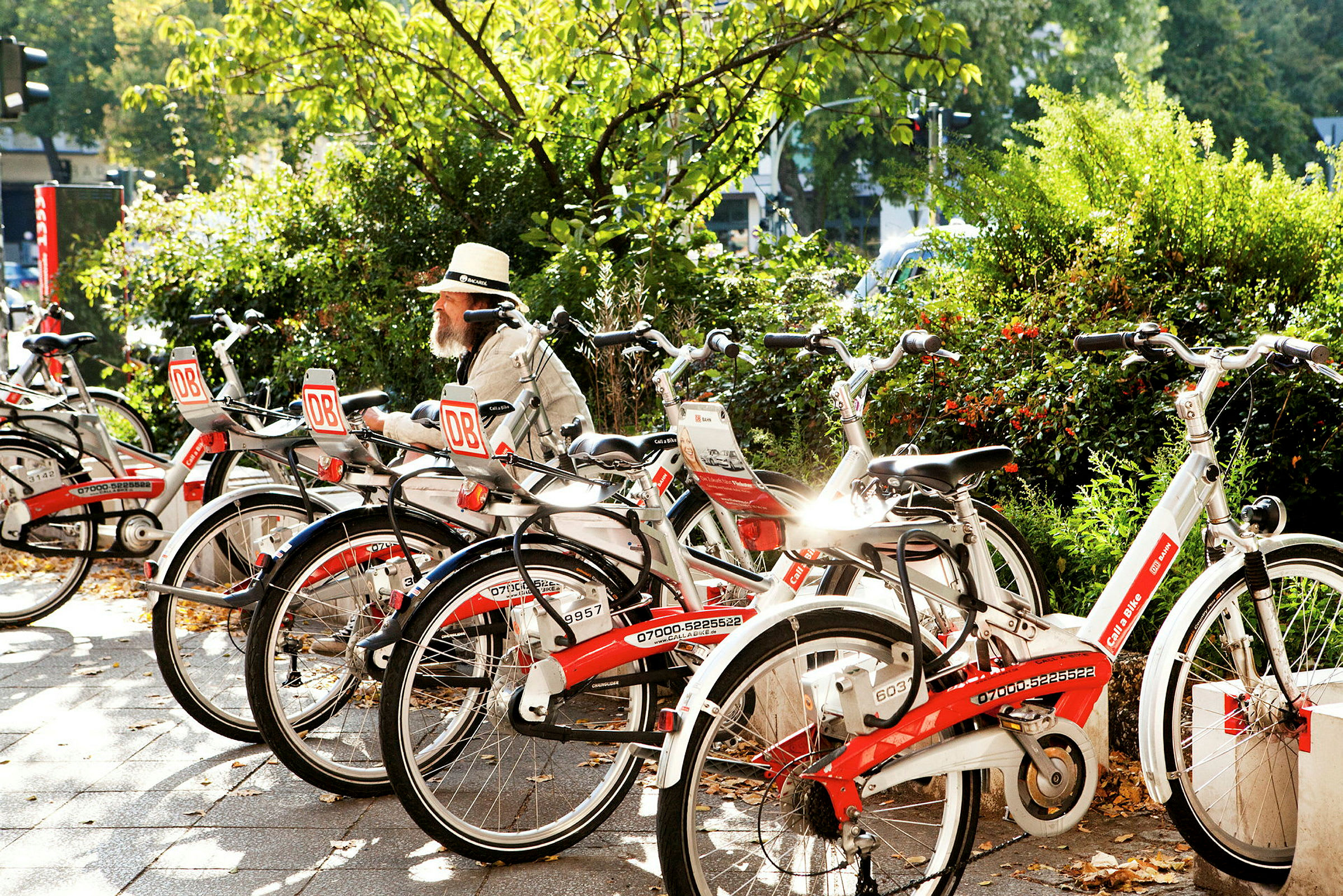 En man med hatt sitter på en bänk omgiven av parkerade cyklar vid en grönskande plats. (AI-genererad text)