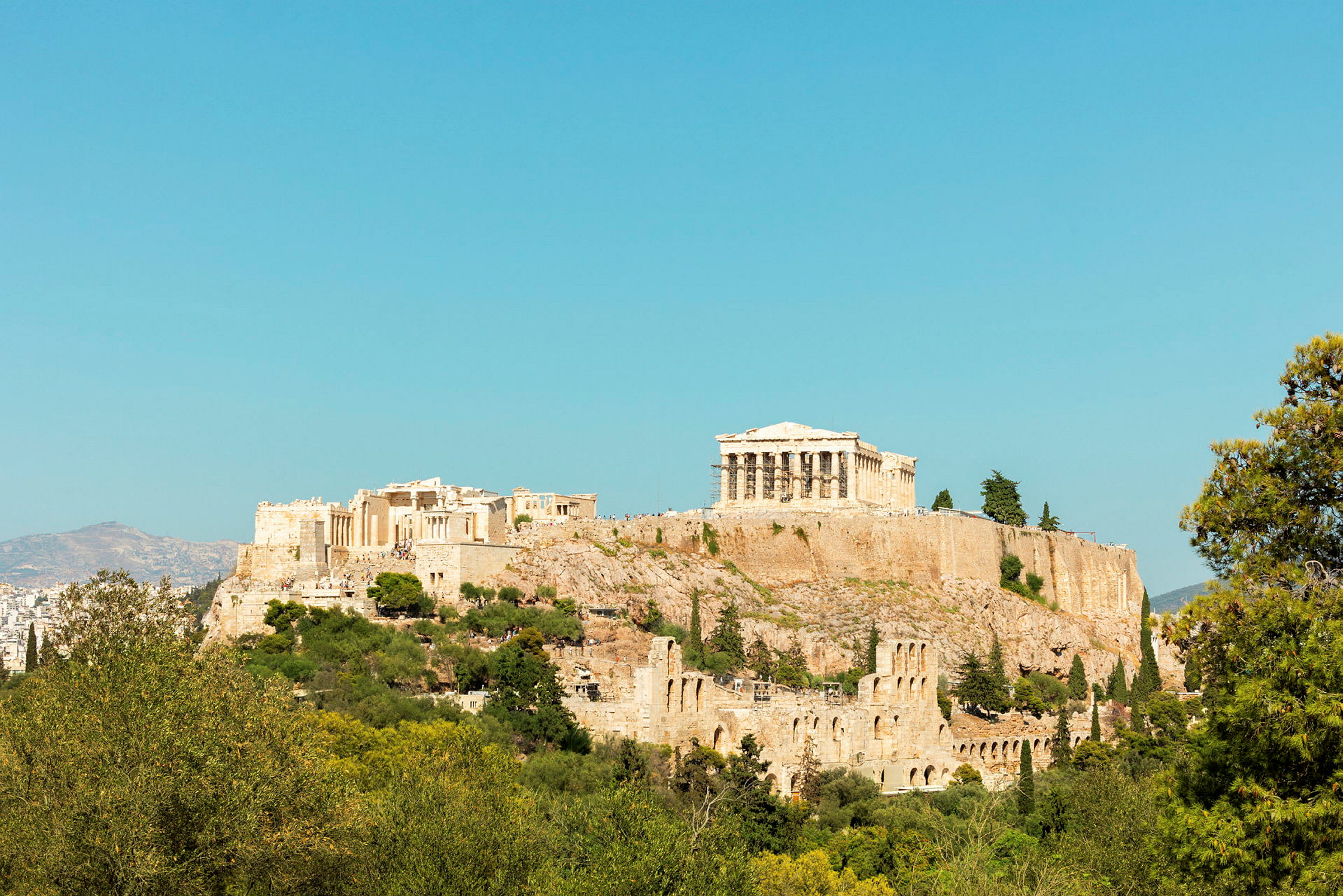 Fotografi av Akropolis med Parthenon på toppen, omgiven av grönska och med klarblå himmel i bakgrunden. (AI-genererad text)