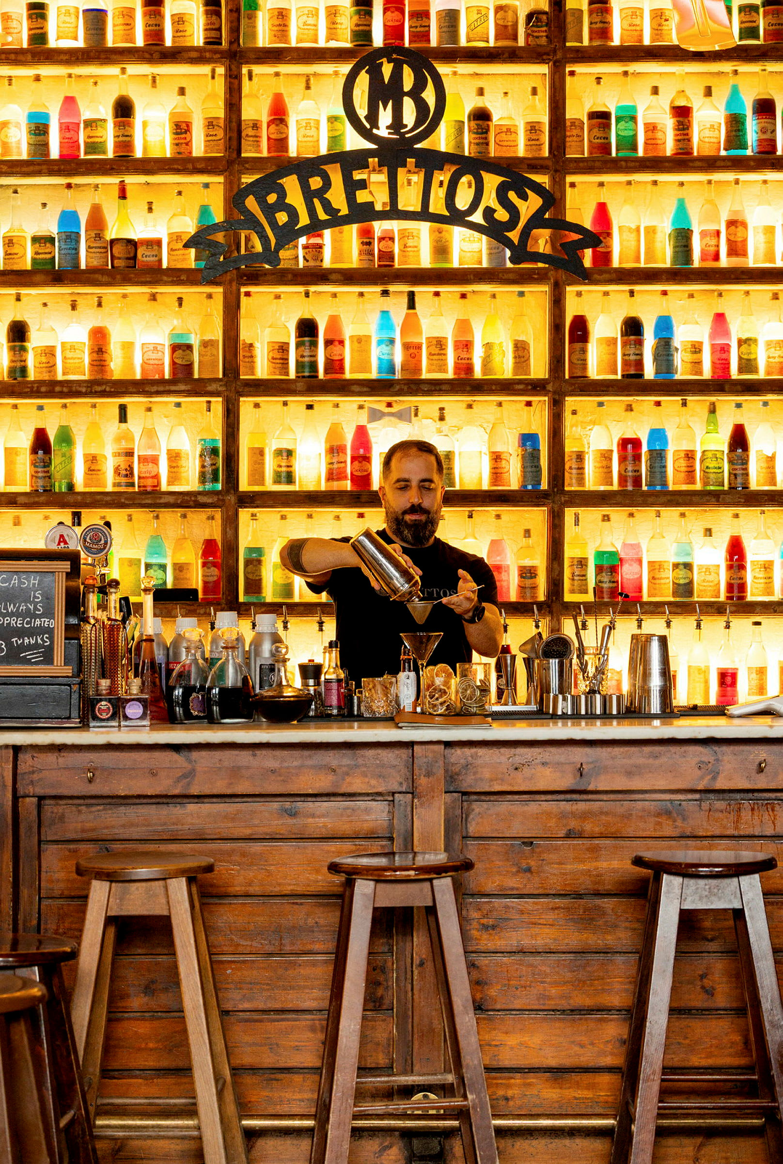 En bar med en vägg full av färgade flaskor, en trädisk och pallar. (AI-genererad text)