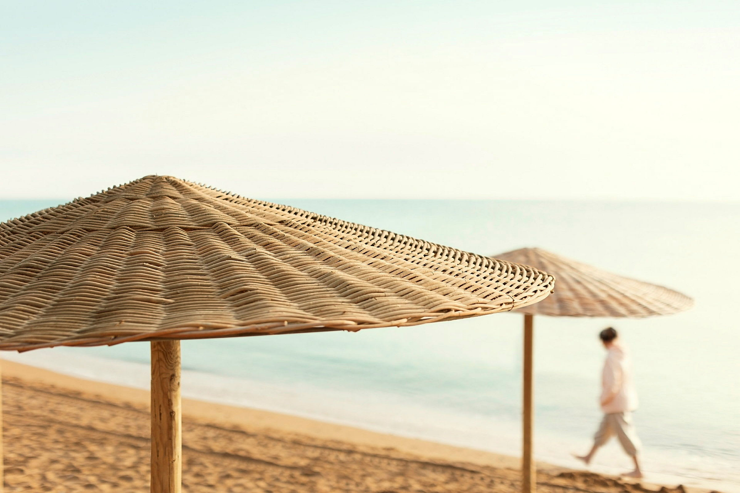 Strand med två solparasoller av rotting och en person som promenerar vid vattnet. (AI-genererad text)