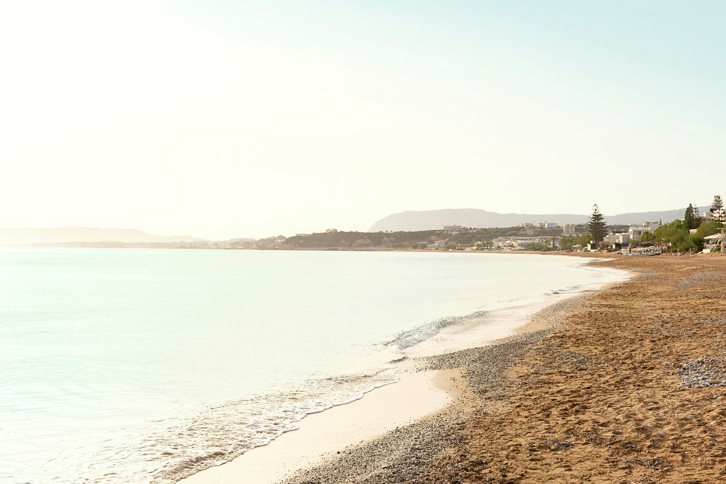 Strand med sand och småsten, med havet vid sidan och byggnader i bakgrunden. (AI-genererad text)