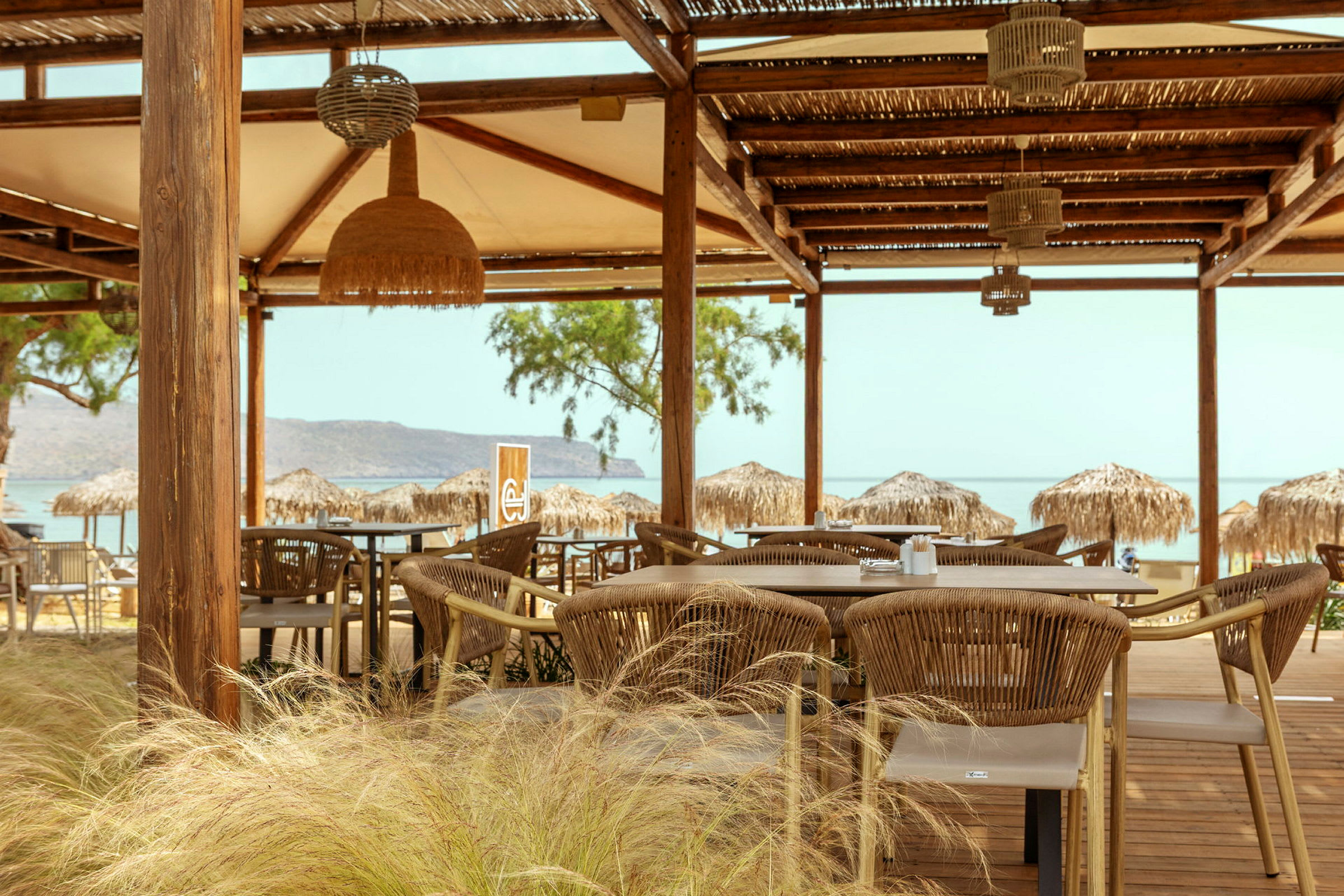 En utomhusrestaurang med bord och stolar under ett träskjul. Bakgrunden visar stranden och havet. (AI-genererad text)