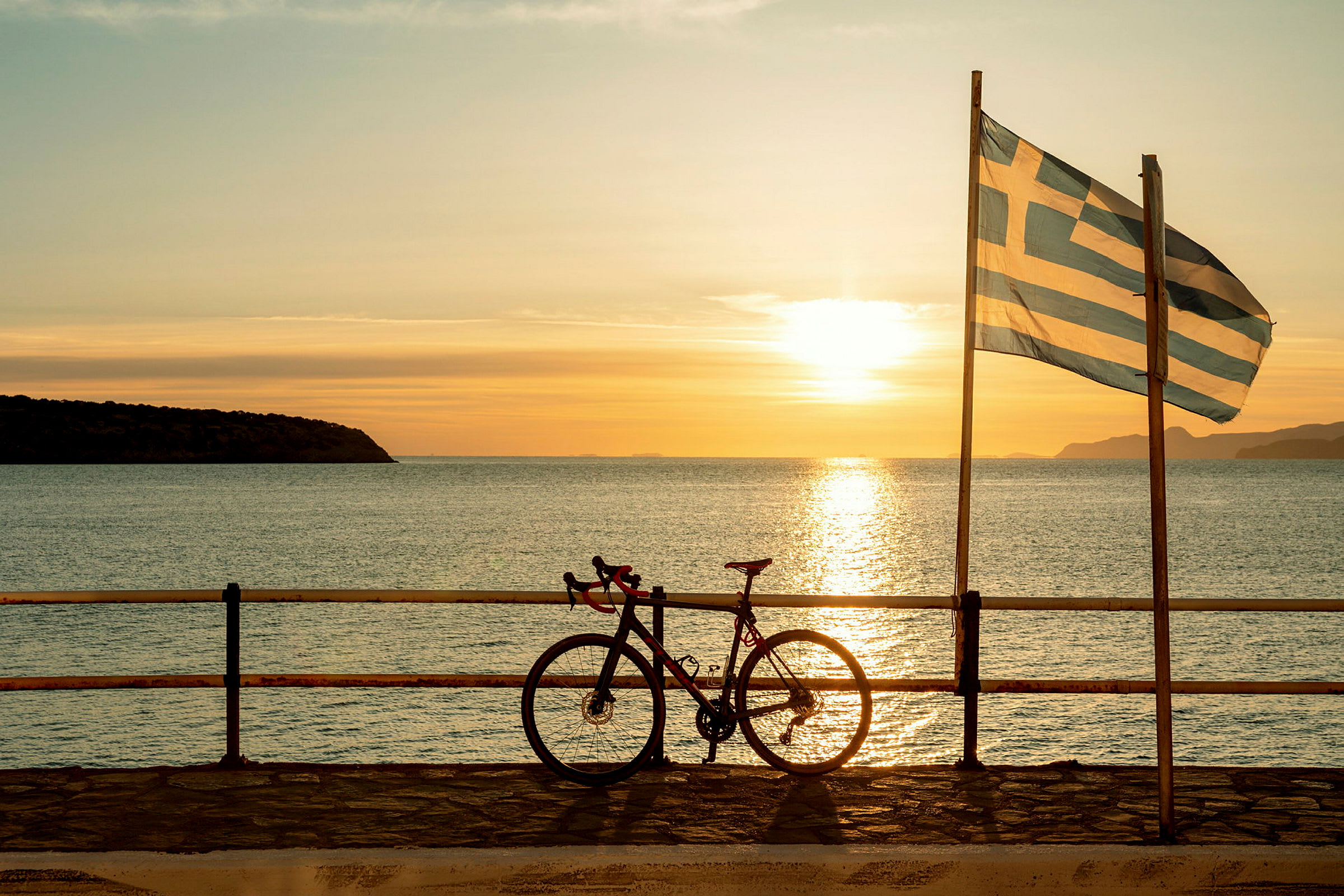 En cykel står lutad mot ett räcke vid havet med en grekisk flagga och en solnedgång i bakgrunden. (AI-genererad text)