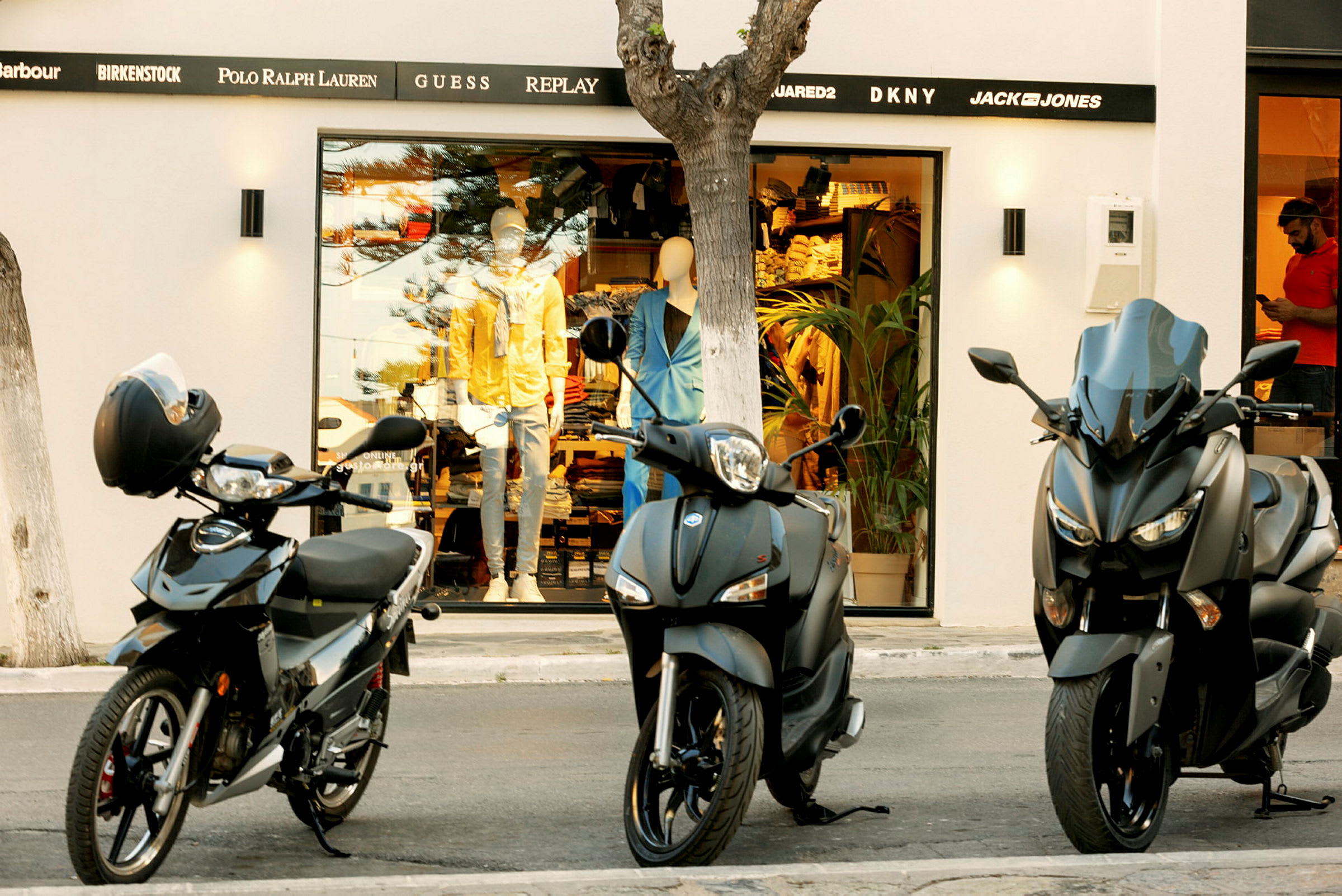 Tre motorcyklar står parkerade framför en butik med skyltfönster och skyltdockor. (AI-genererad text)