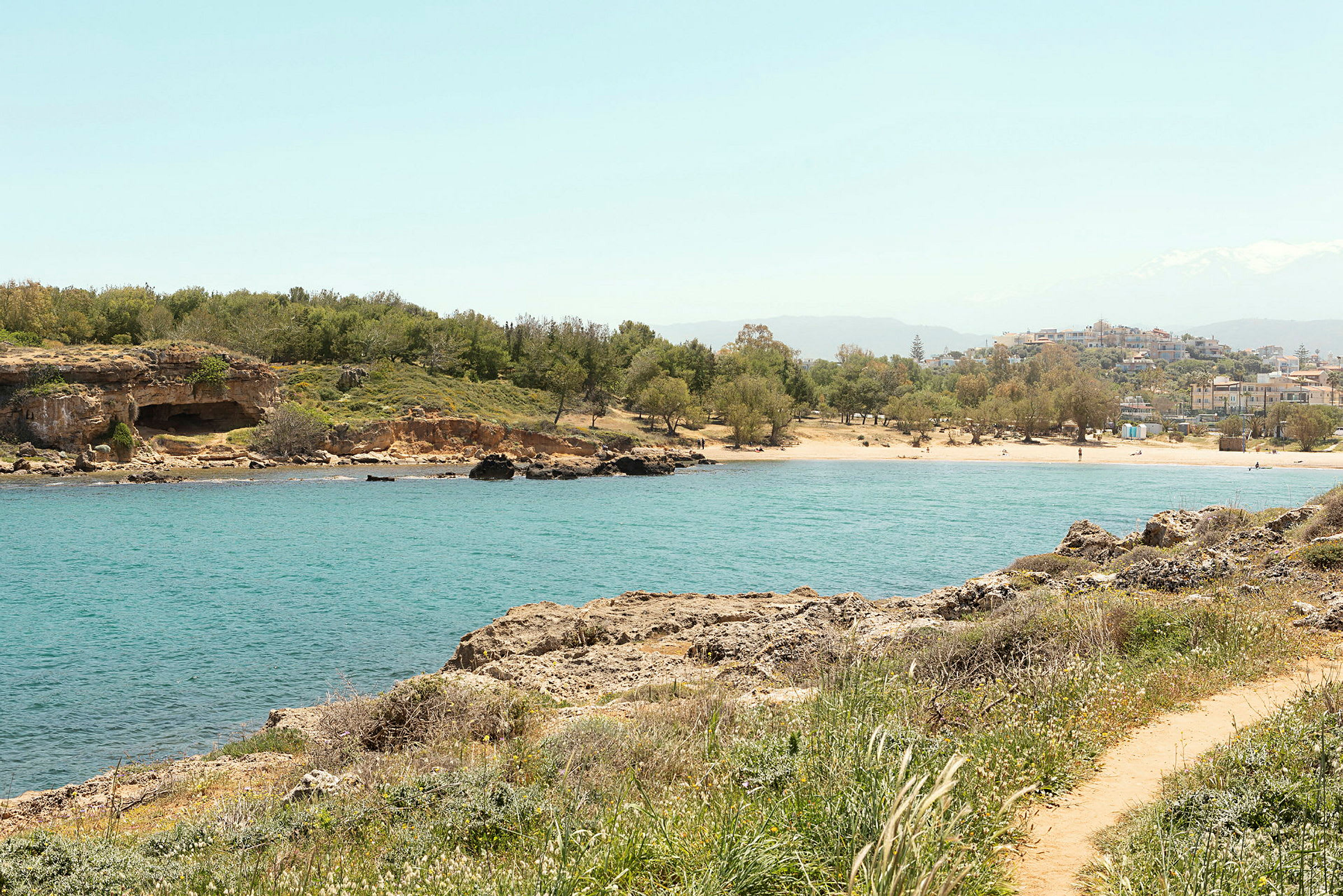 En strand med klart vatten och grönska, med byggnader i bakgrunden. (AI-genererad text)