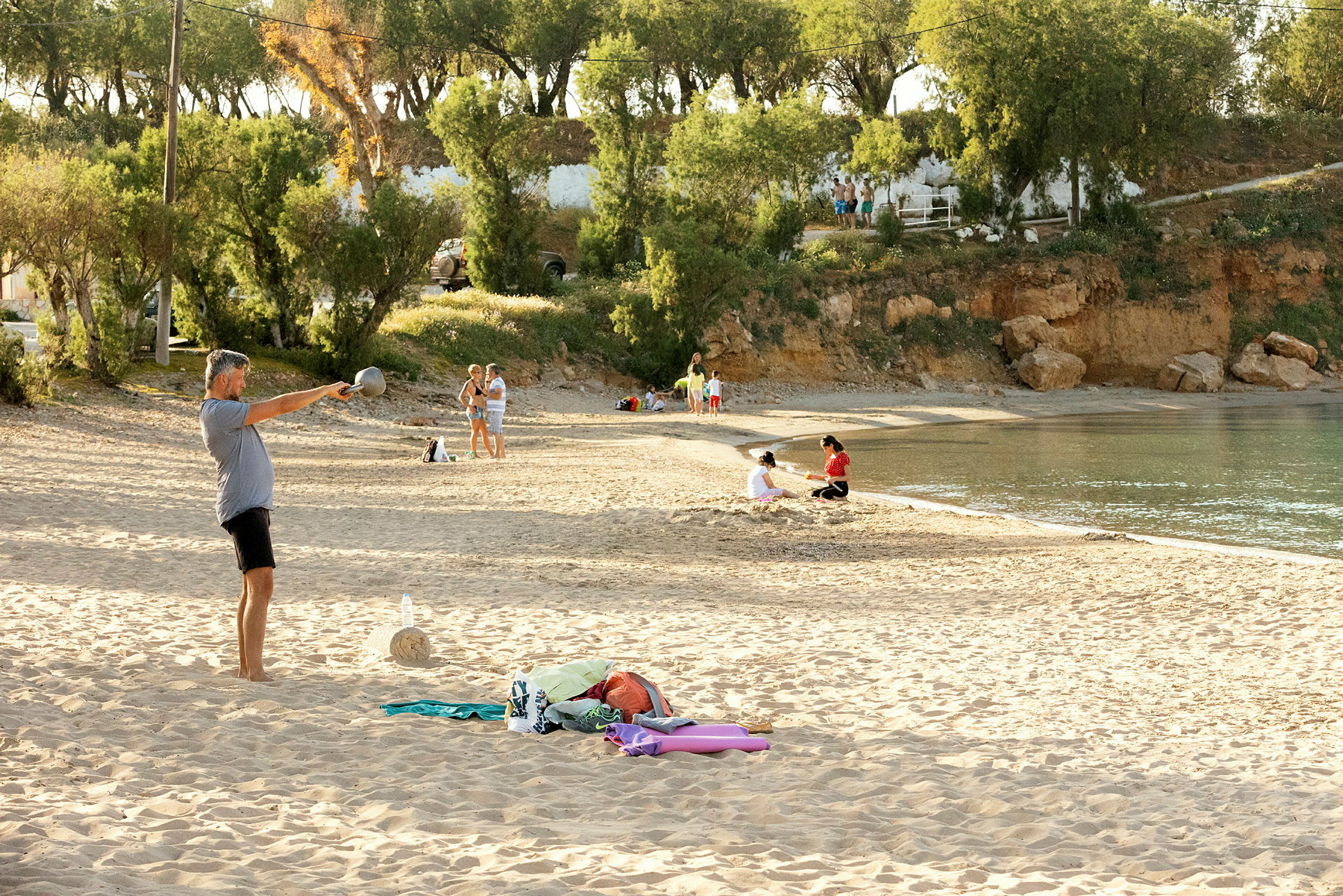 Strand med människor på sanden och vid vattenkanten, omgiven av träd och klippor. (AI-genererad text)