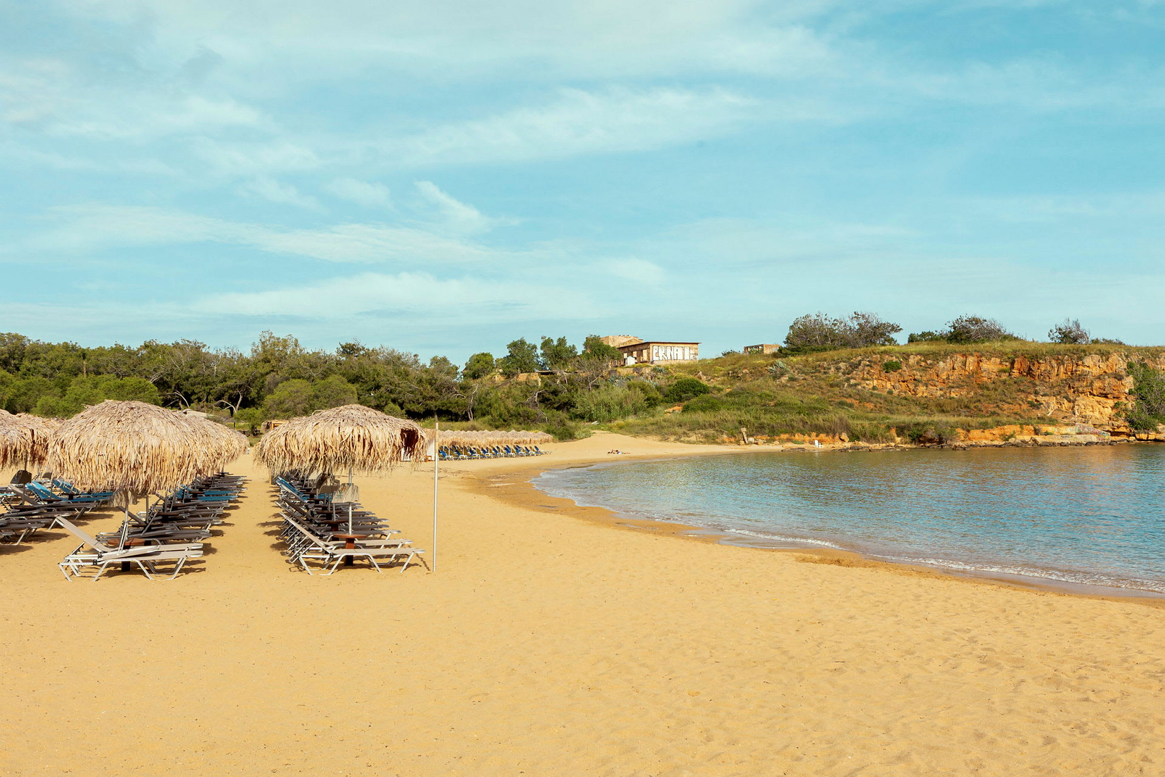 En sandstrand med solstolar och parasoller gjorda av halm, omgiven av grönska och havet syns till höger. (AI-genererad text)