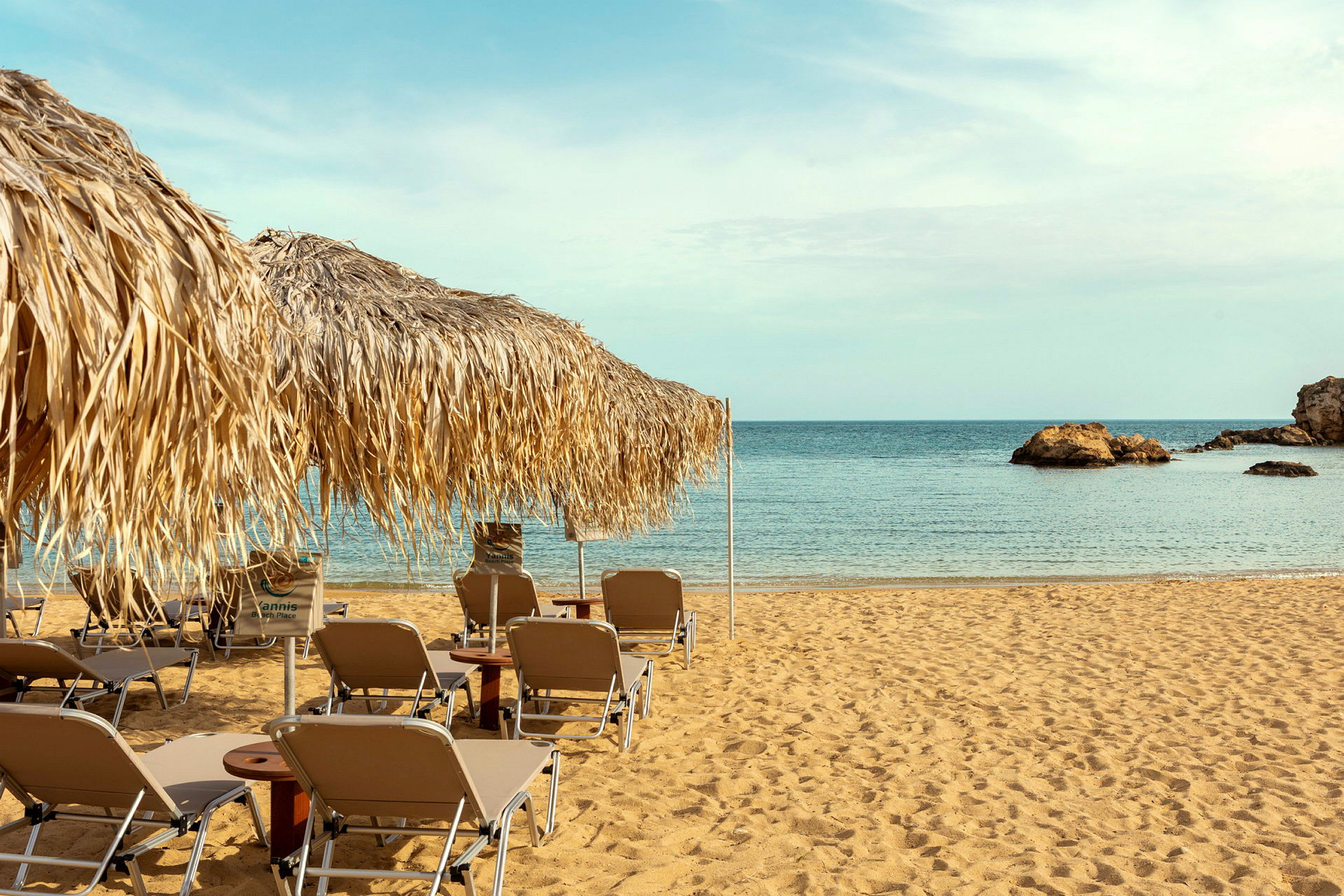 Strand med solstolar och parasoller av halm, med utsikt över havet och klippformationer. (AI-genererad text)