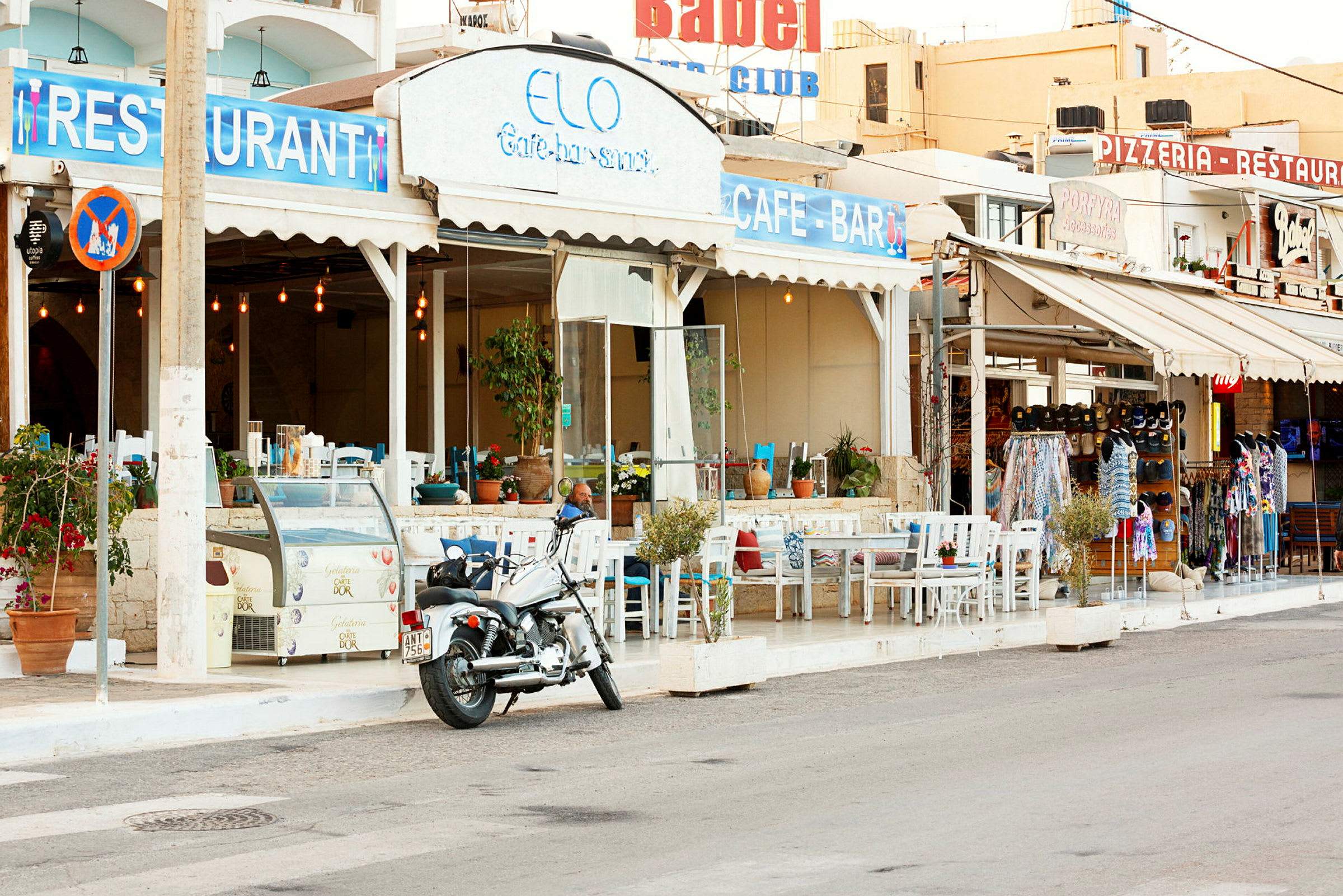 Motiv av ett café med uteservering vid en gata, ett skylt med texten "Restaurang" och en motorcykel parkerad i förgrunden. (AI-genererad text)