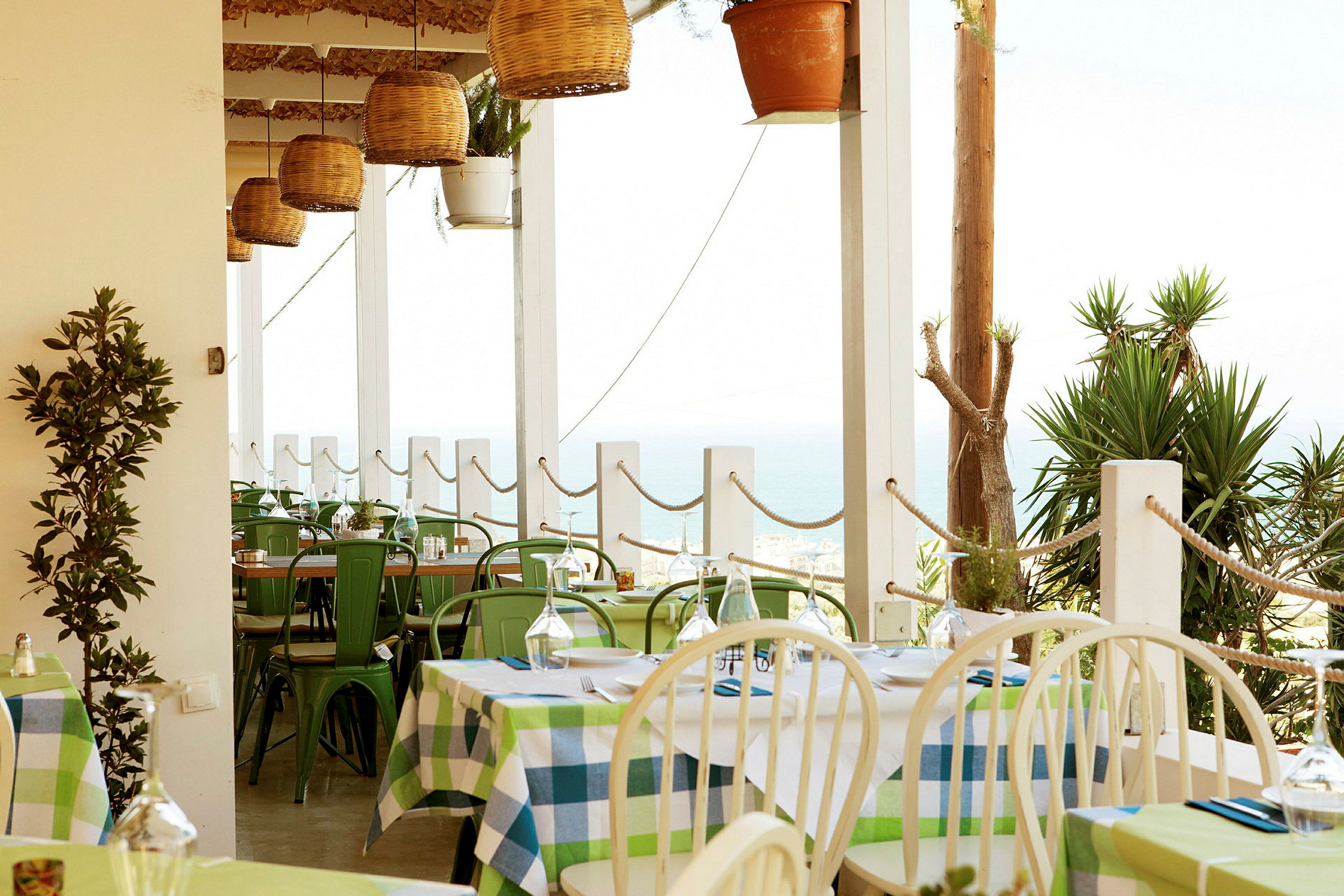 En utomhusrestaurang med färgglada dukar, gröna stolar och utsikt över havet. (AI-genererad text)