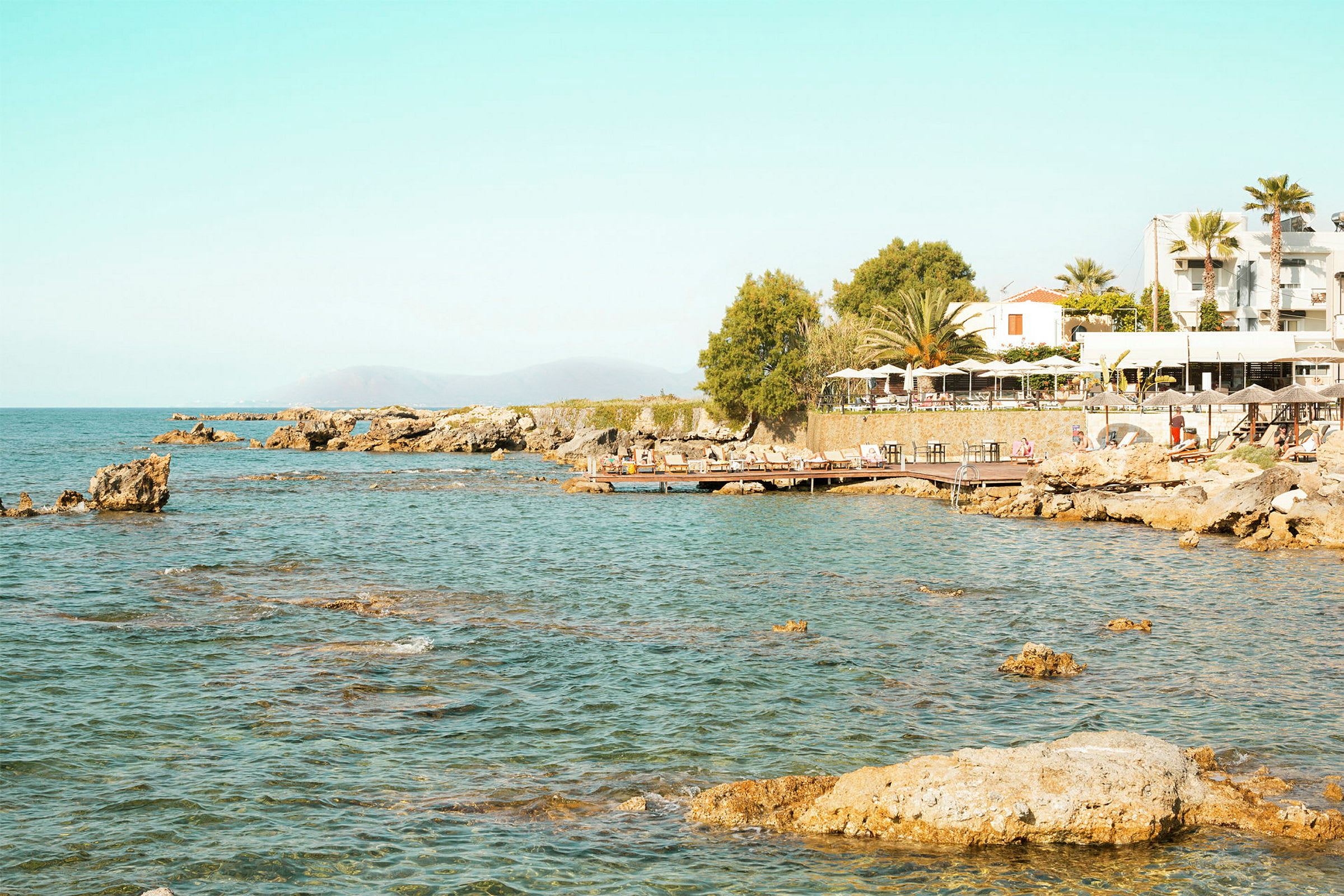 Rocks and water with a building and sun loungers to the right. (AI-genererad text)