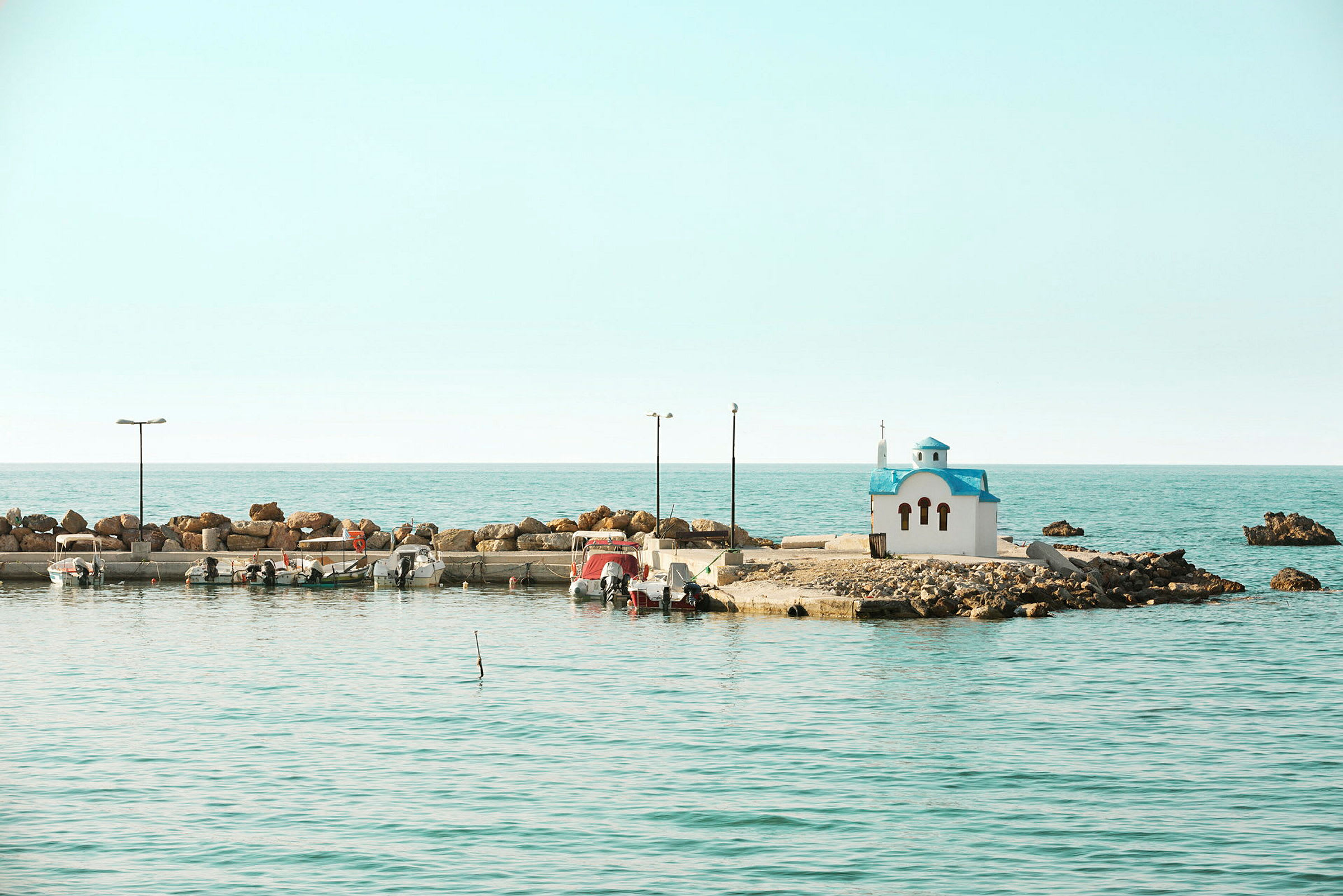 Vit byggnad med blått tak vid havet, liten hamn med båtar. (AI-genererad text)