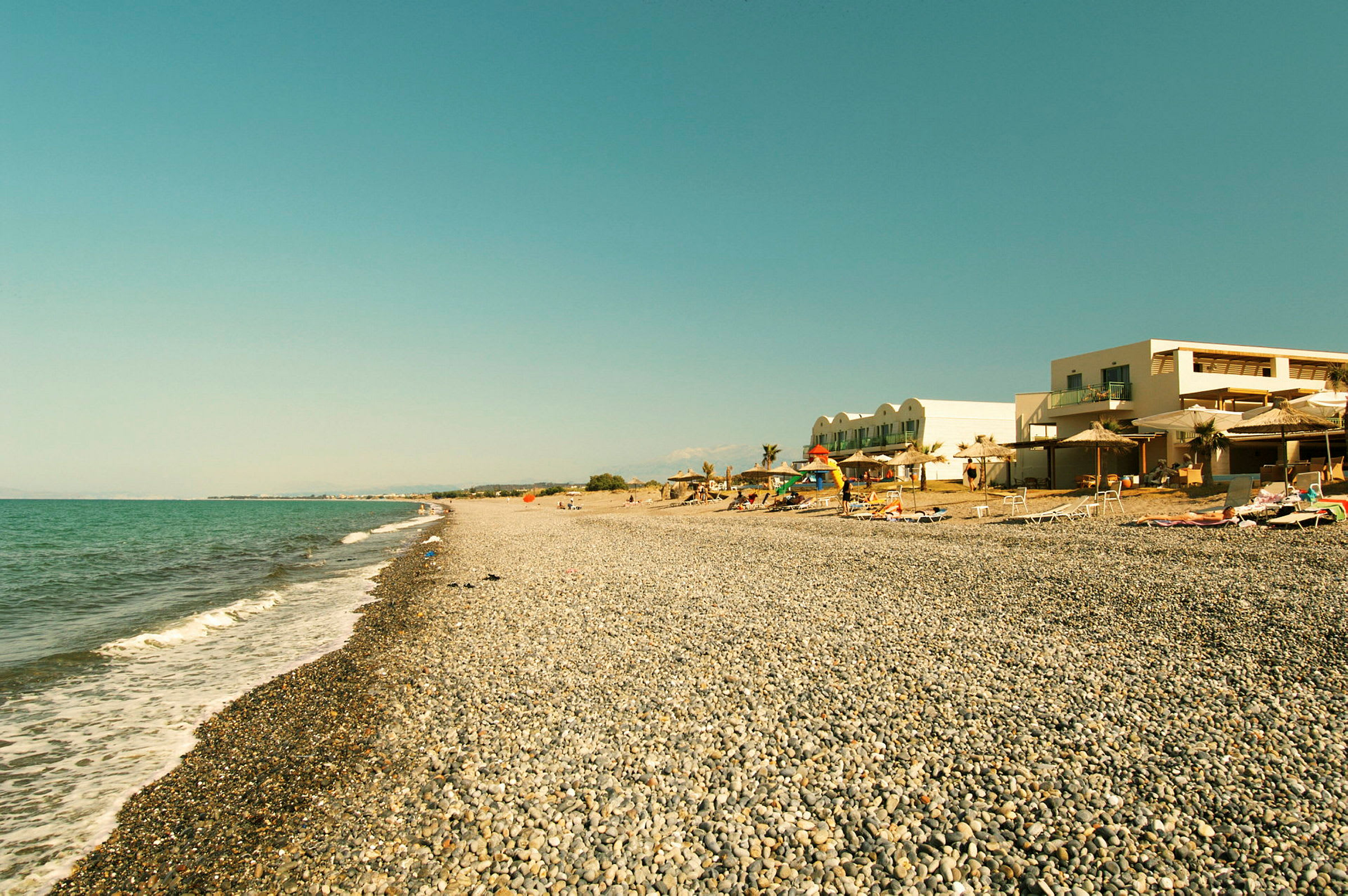 Strand med runda stenar och hotellbyggnad i bakgrunden. (AI-genererad text)