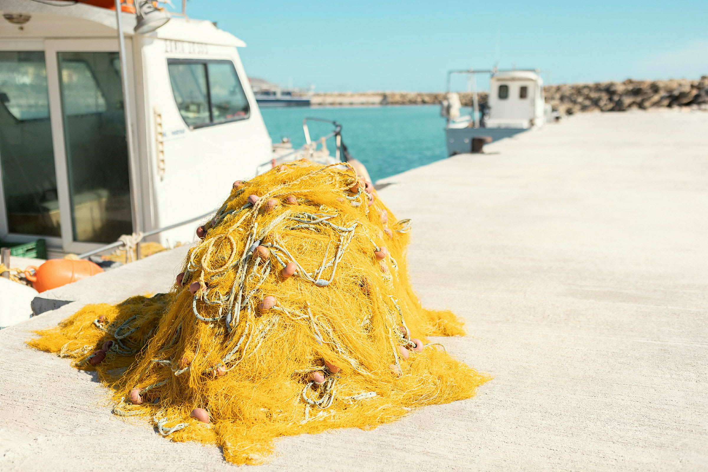 En hög med gult fisknät ligger på en kaj, med båtar och havet i bakgrunden. (AI-genererad text)