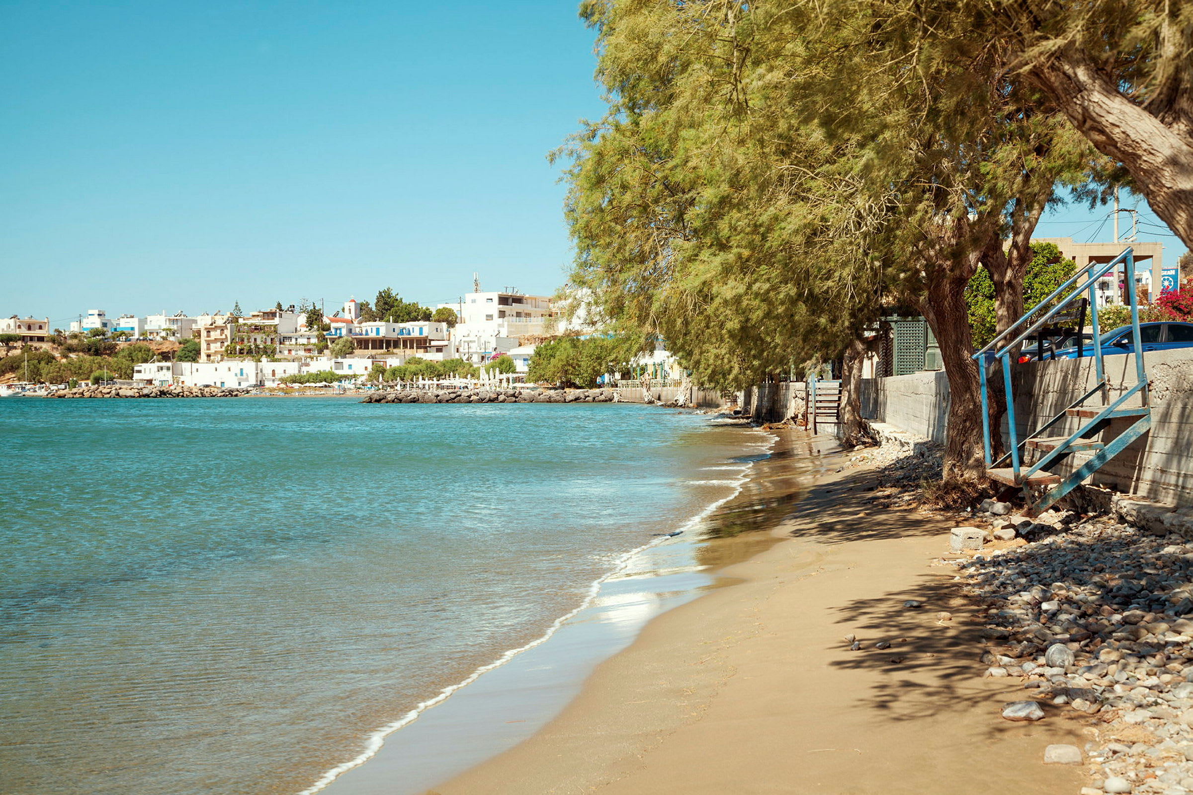 Strand med sand och sten, träd och steg, med en liten stad och hav i bakgrunden. (AI-genererad text)