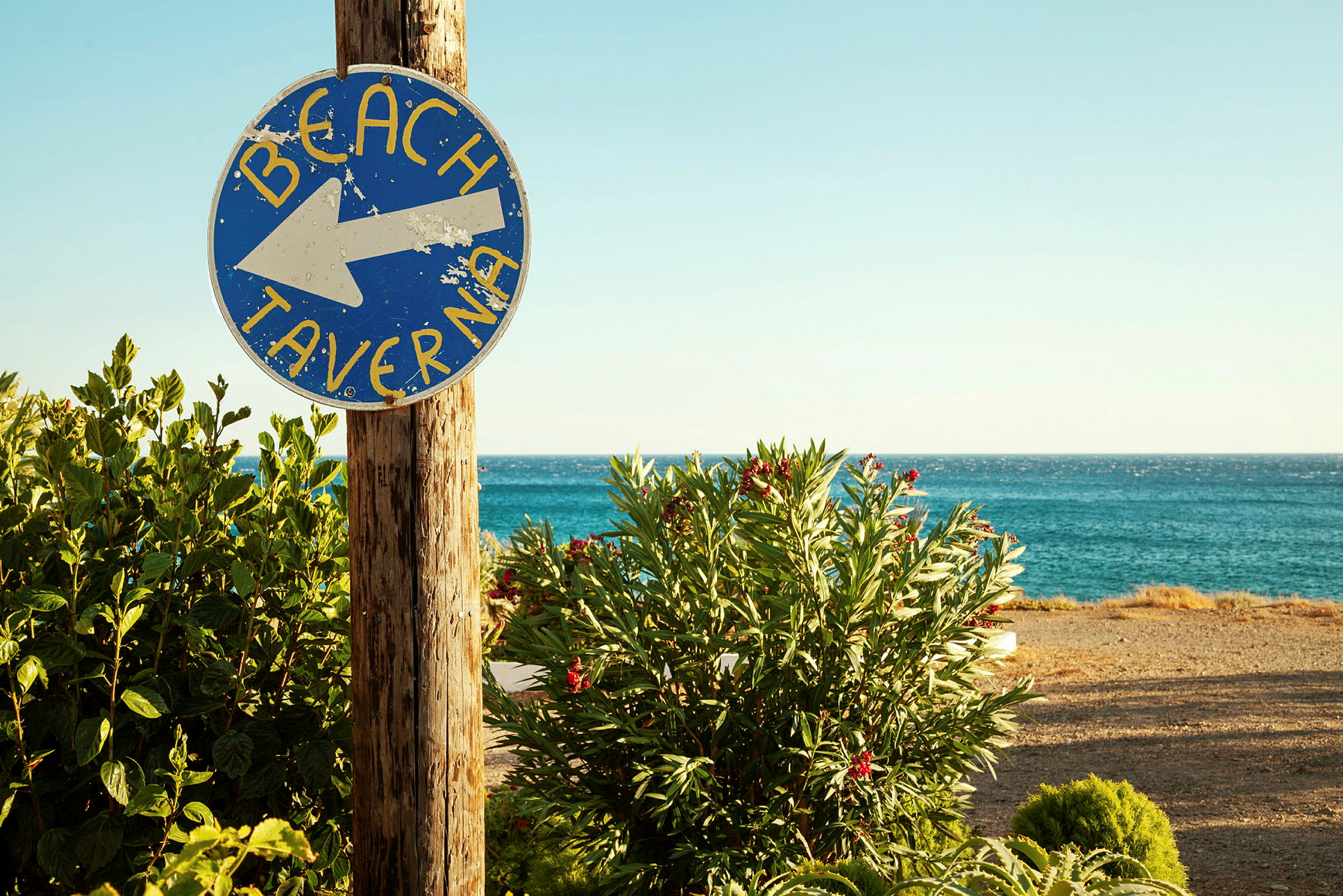 En skylt med texten 'Beach Taverna' pekar mot stranden, med grönska och havet i bakgrunden. (AI-genererad text)