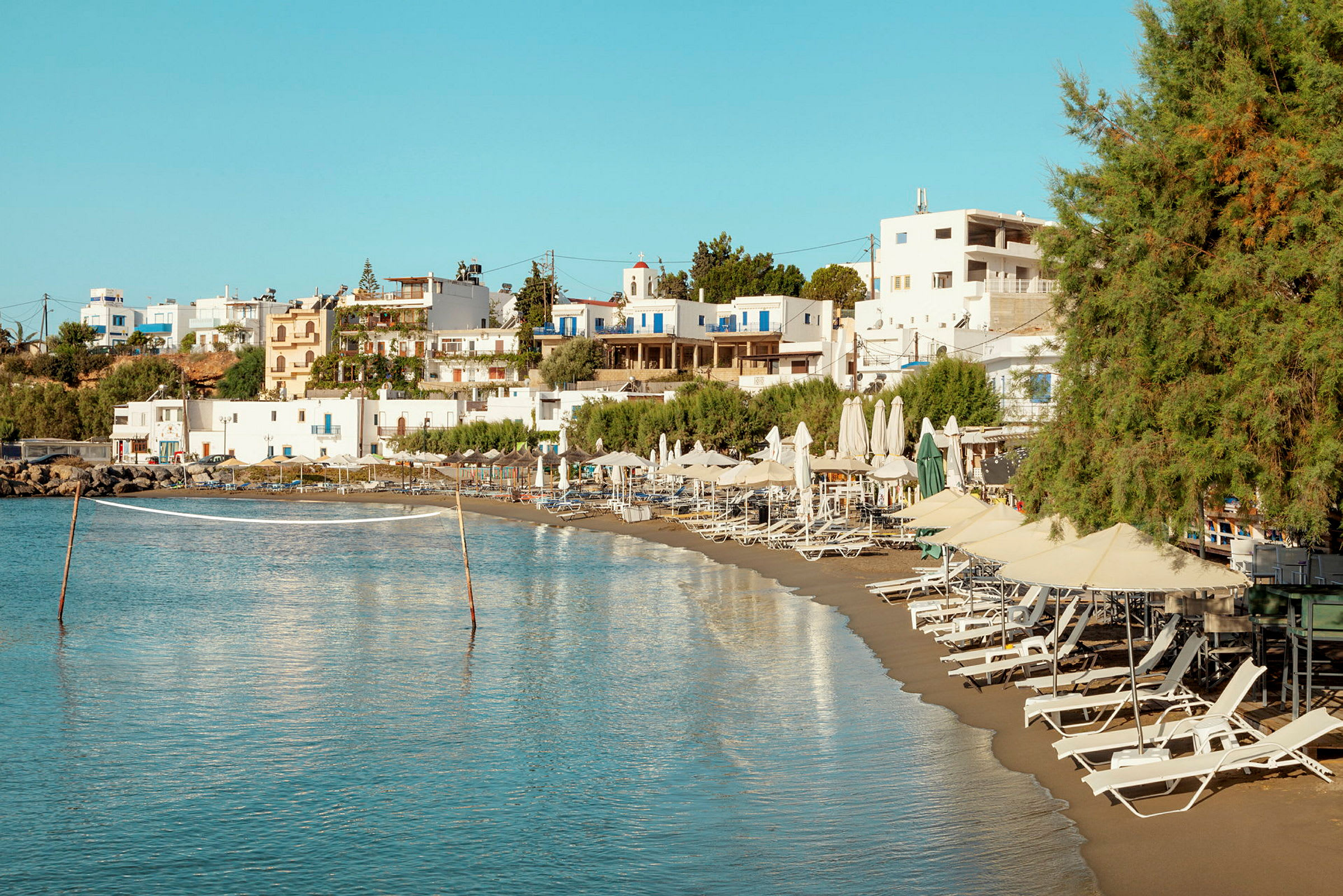 Strand med solstolar och parasoller samt vitmålade byggnader i bakgrunden. (AI-genererad text)