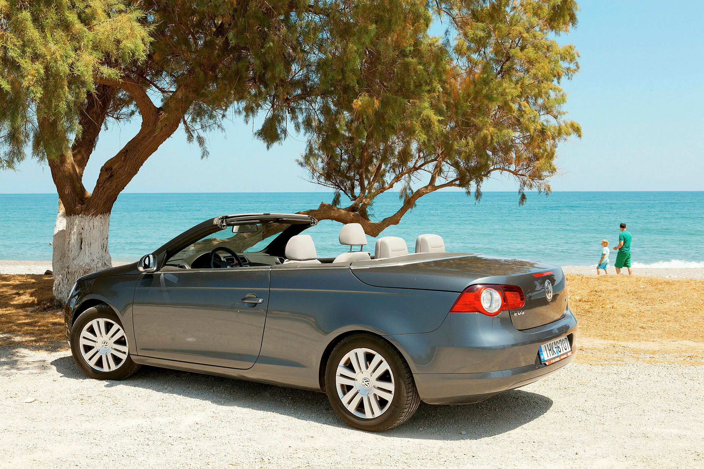 En grå cabriolet står parkerad vid en strand med hav och träd i bakgrunden. (AI-genererad text)
