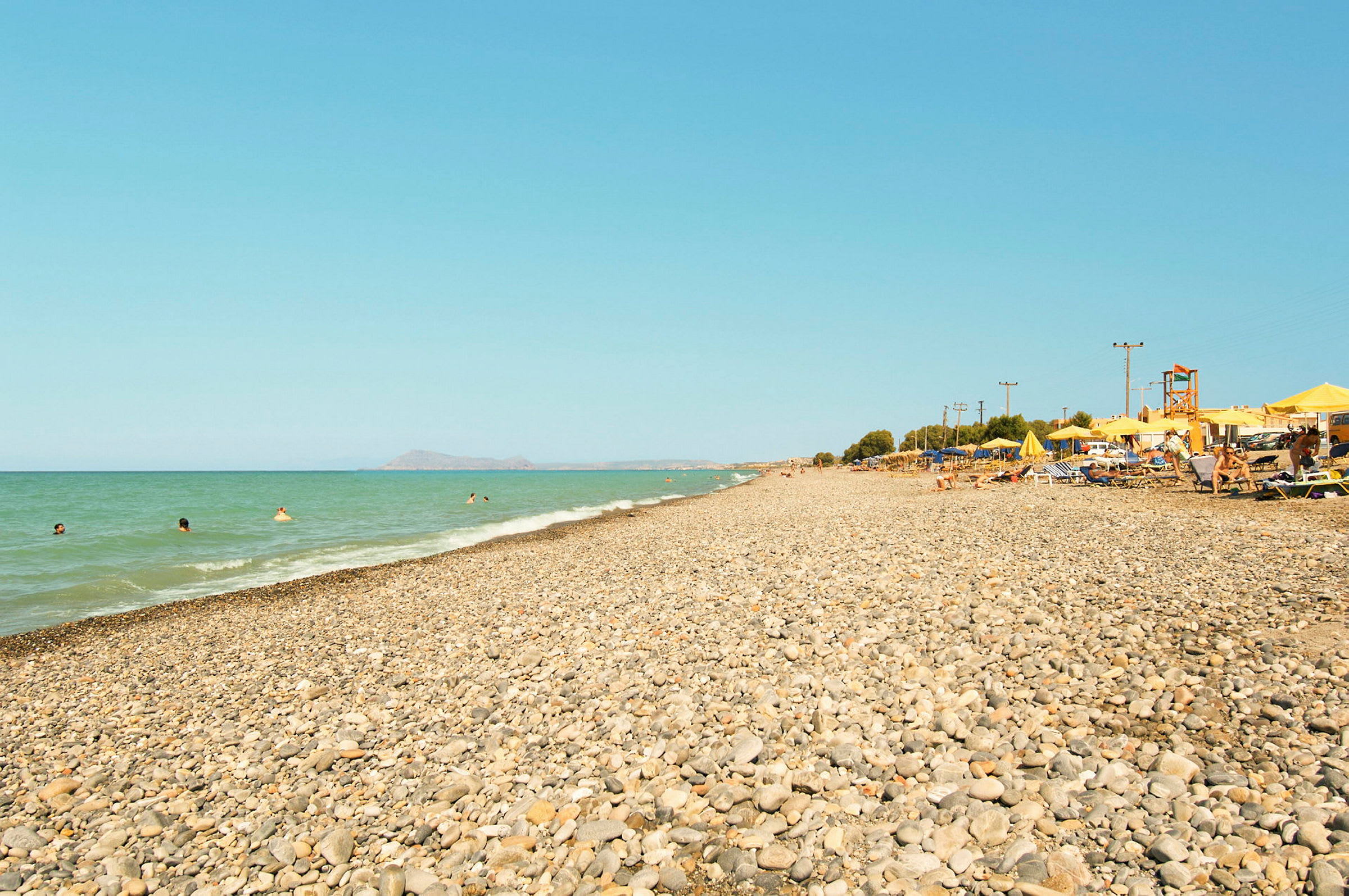 En klapperstensstrand med havet i bakgrunden och parasoller och solstolar till höger. (AI-genererad text)