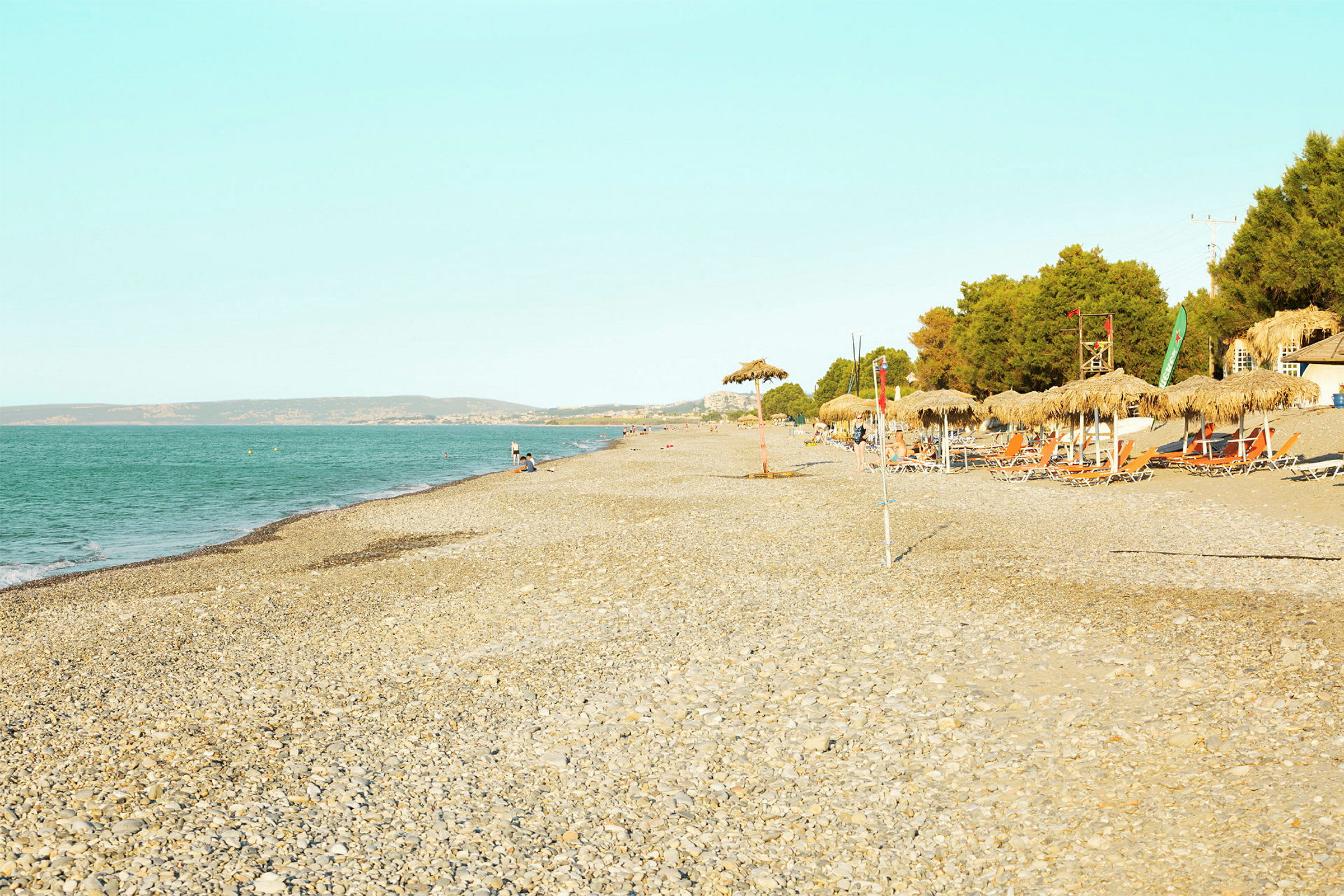 En strand med sten, parasoller och solstolar bredvid en skog. Havet är synligt till vänster. (AI-genererad text)