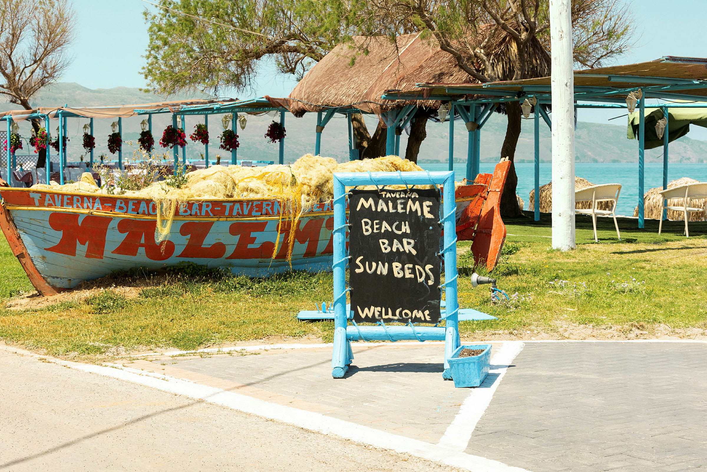 Skylt med texten 'Maleme Beach Bar Sunbeds Welcome' framför en båt dekorerad med nät samt uteservering vid havet. (AI-genererad text)