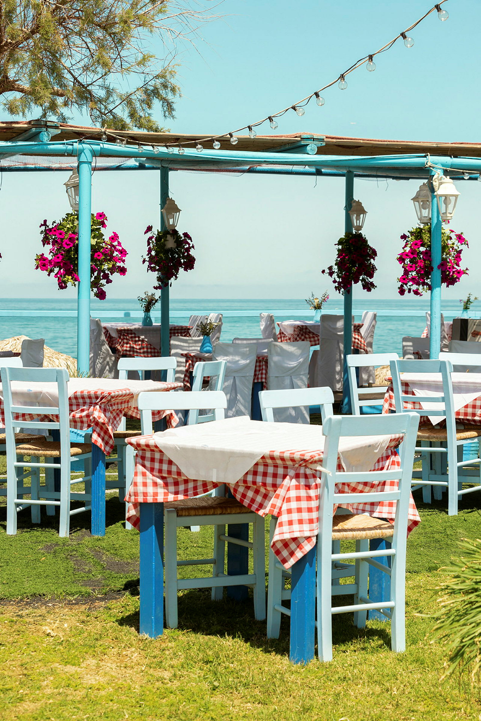 Bild av tomma restaurangbord med vita och rödrutiga dukar, blå stolar och blomkrukor på gräs nära havet. (AI-genererad text)