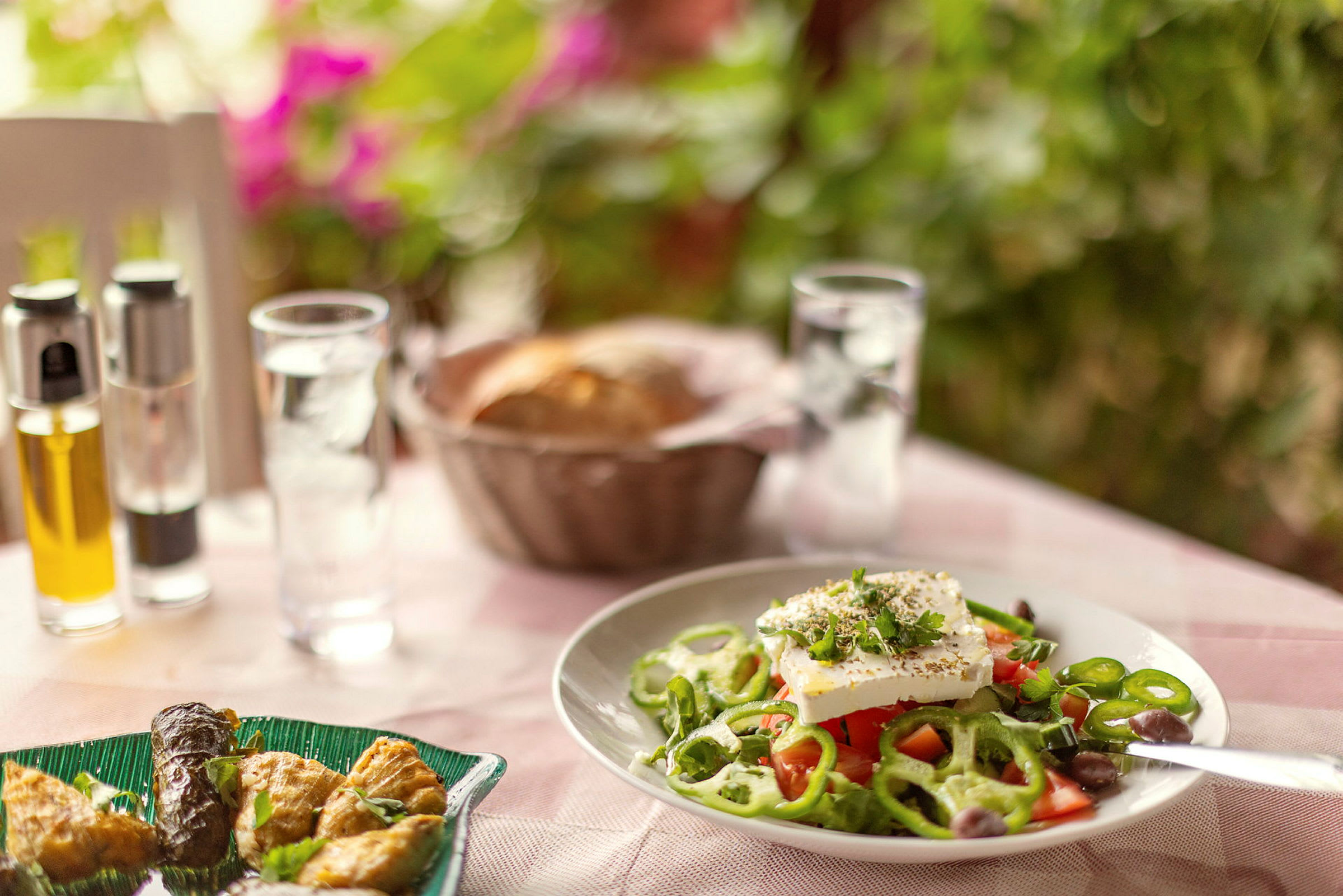 En tallrik med grekisk sallad på ett bord, med olja, peppar och brödkorg i bakgrunden. (AI-genererad text)