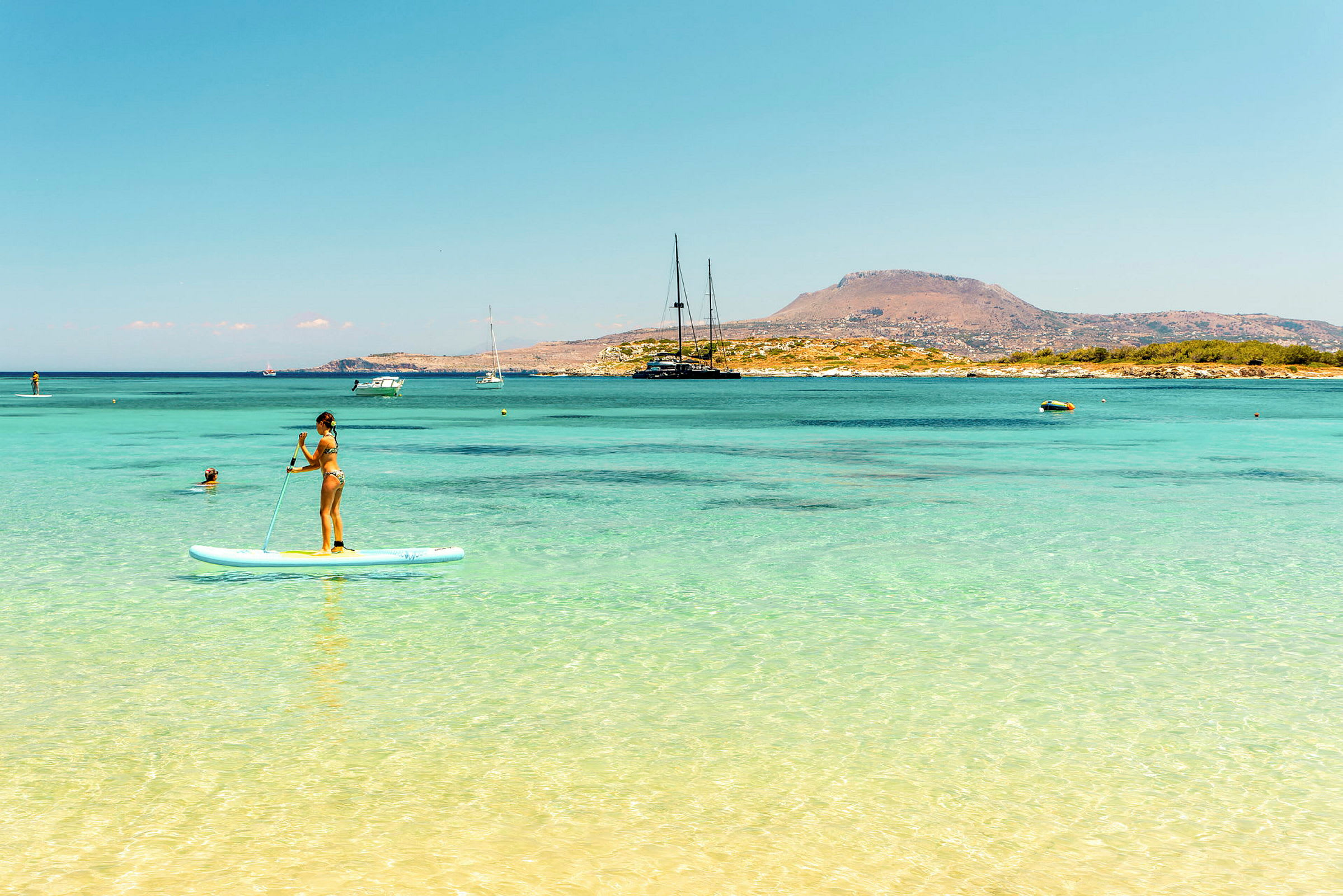 Person som står på en paddleboard i klart blått vatten med en bergig kustlinje i bakgrunden. (AI-genererad text)