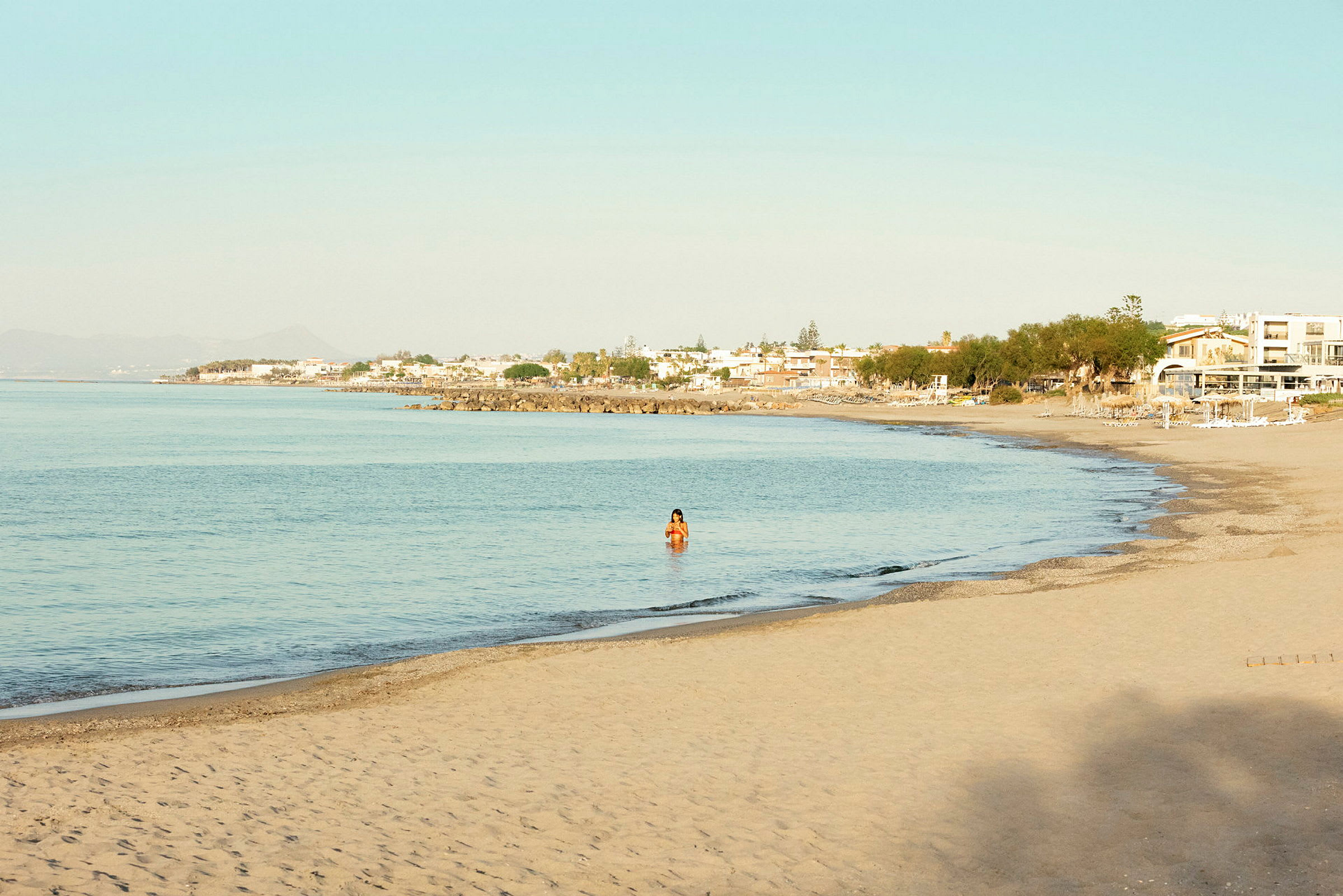 En sandstrand med lugnt blått vatten, en person syns i vattnet och hus i bakgrunden. (AI-genererad text)
