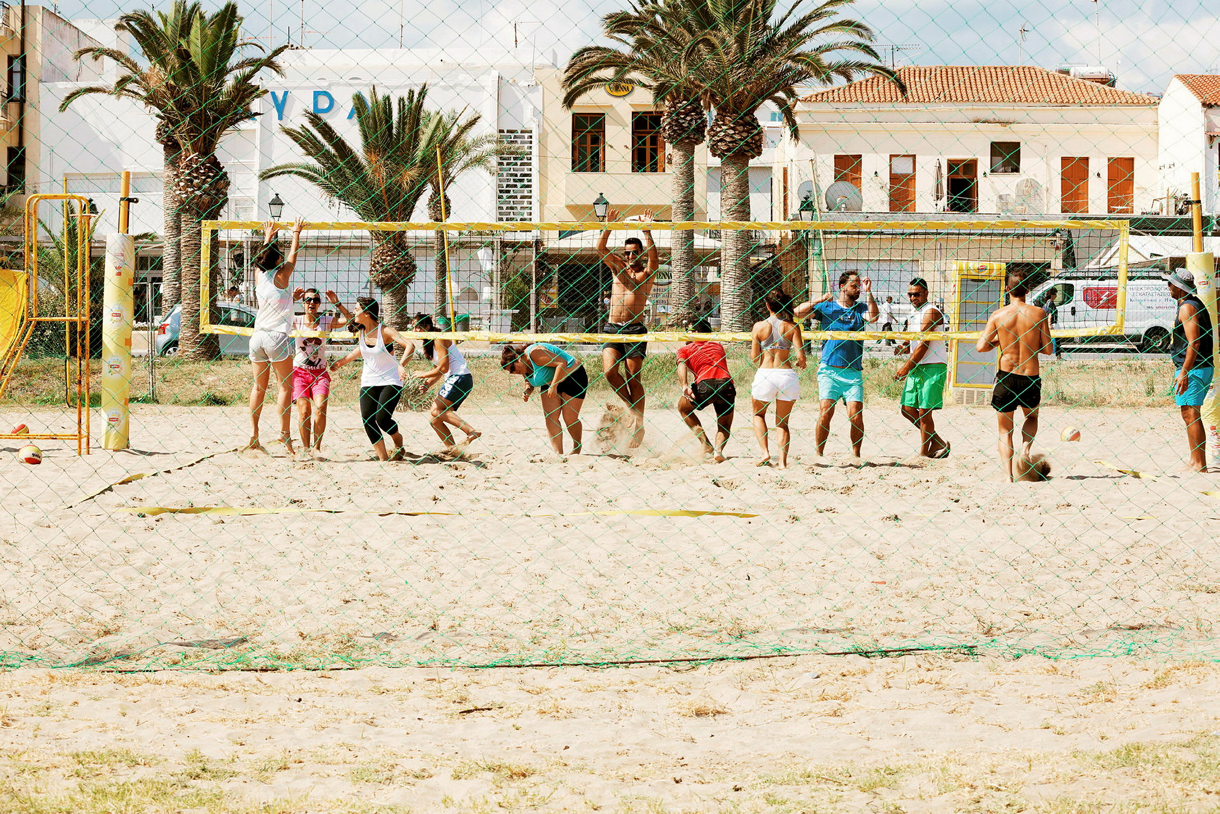 En grupp människor spelar beachvolleyboll på en sandplan framför byggnader och palmer. (AI-genererad text)