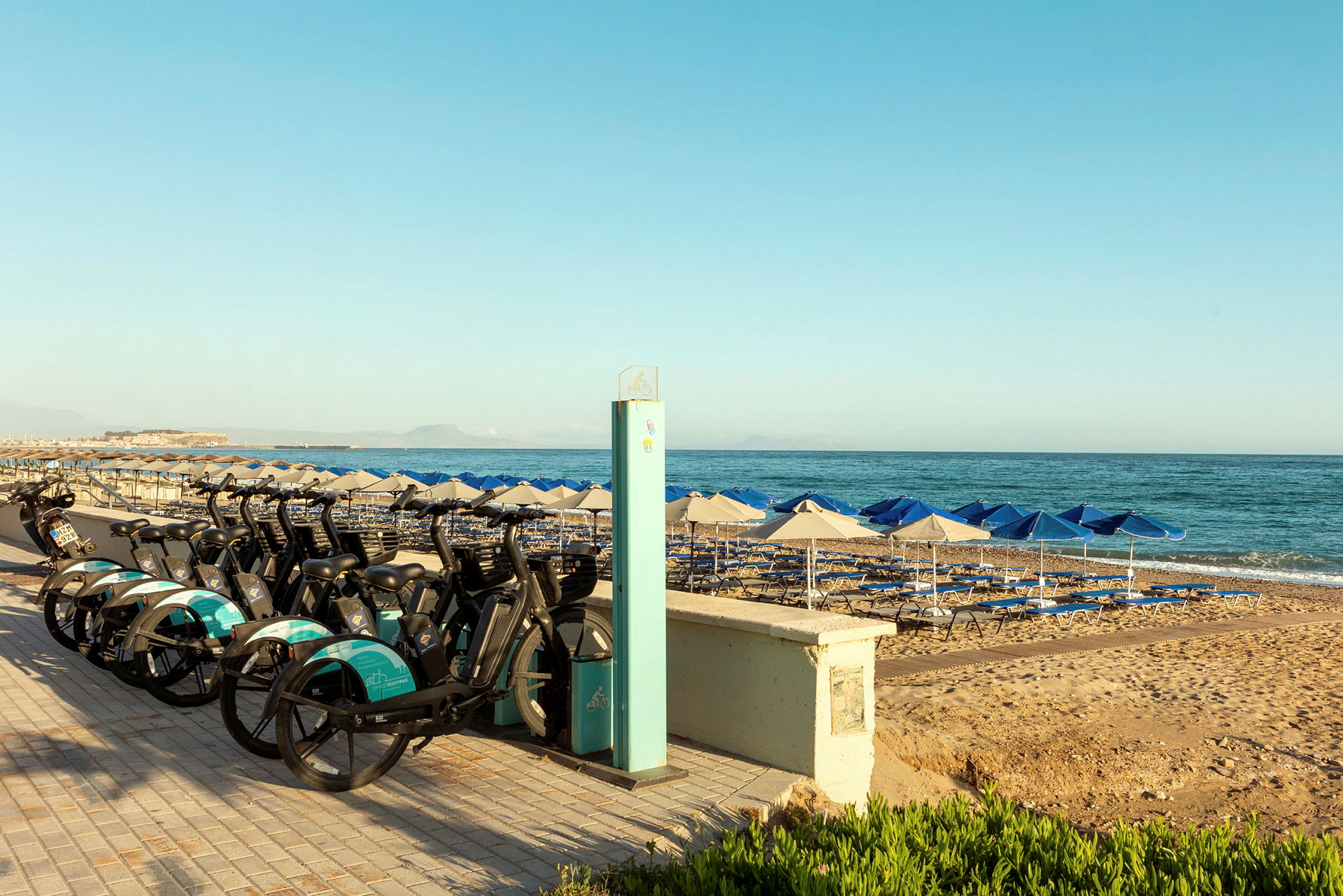 Cyklar parkerade vid en strandpromenad med parasoller och solstolar vid havet. (AI-genererad text)