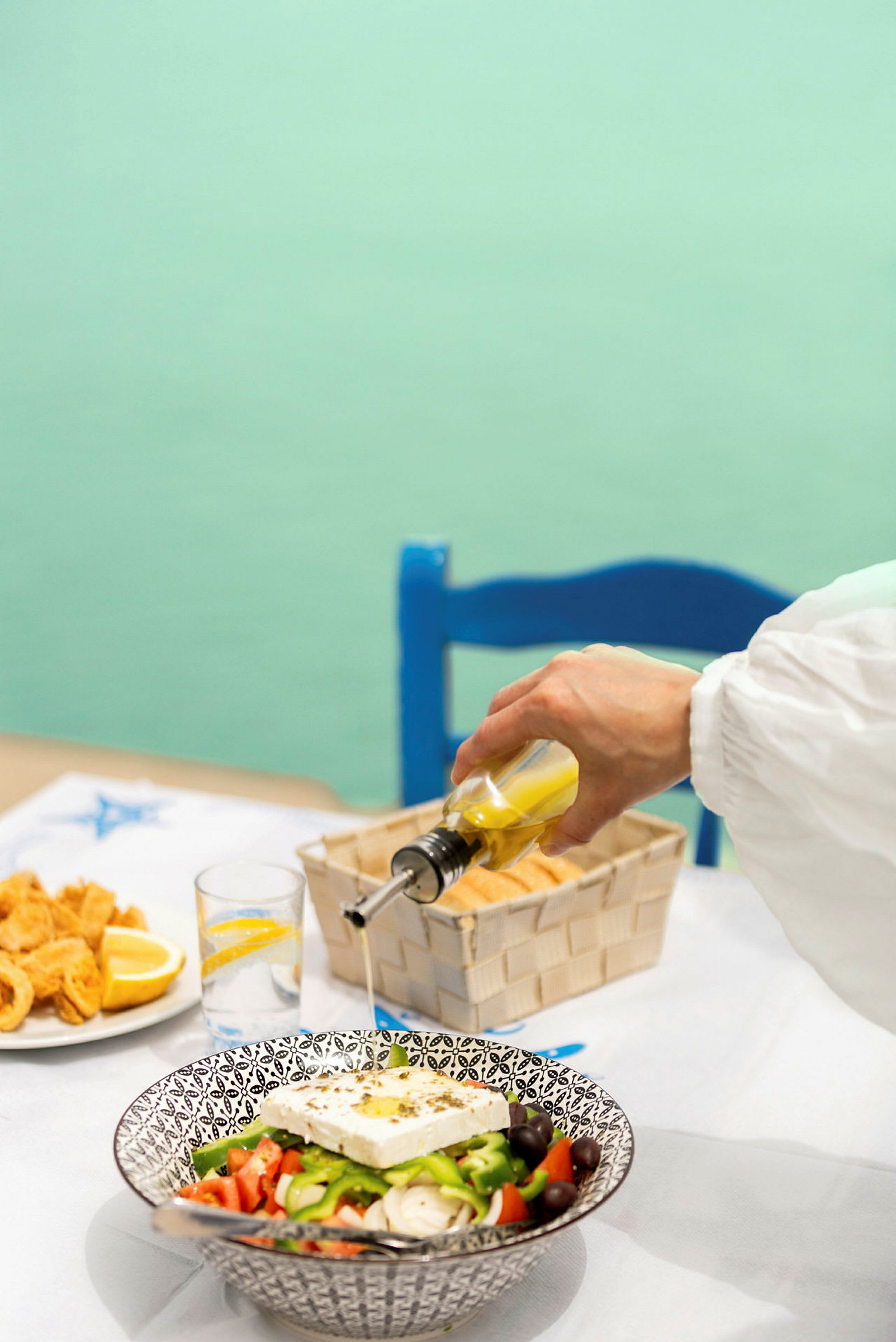 En hand häller olivolja över en grekisk sallad på ett bord. (AI-genererad text)