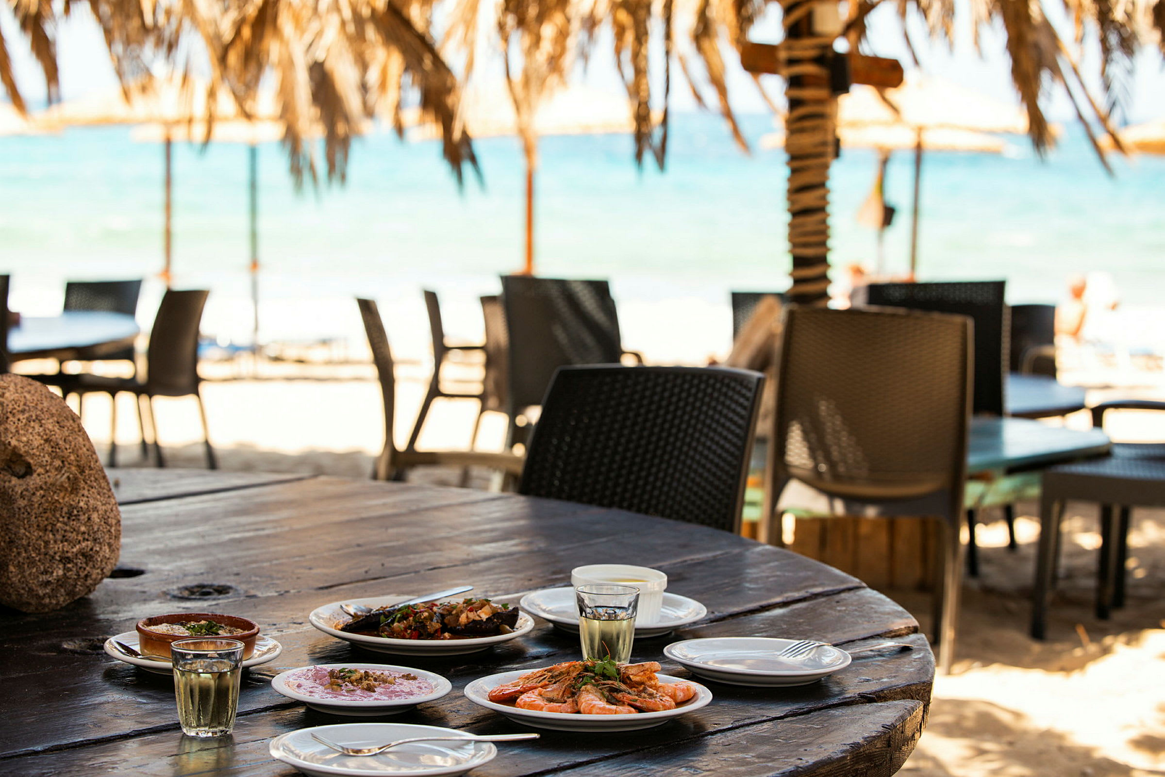 Ett bord med tallrikar fyllda med olika maträtter, med stranden och havet i bakgrunden. (AI-genererad text)