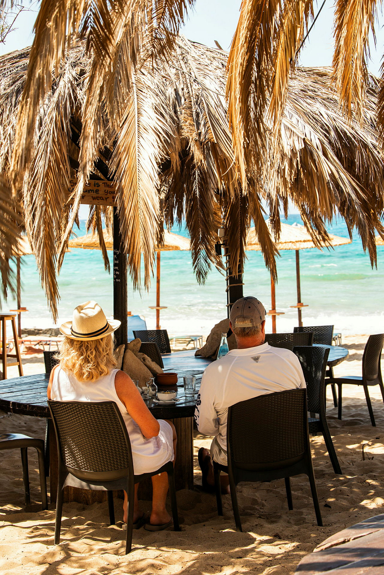 Två personer sitter vid ett bord under en takstruktur med palmblad, med utsikt över en strand och havet. (AI-genererad text)